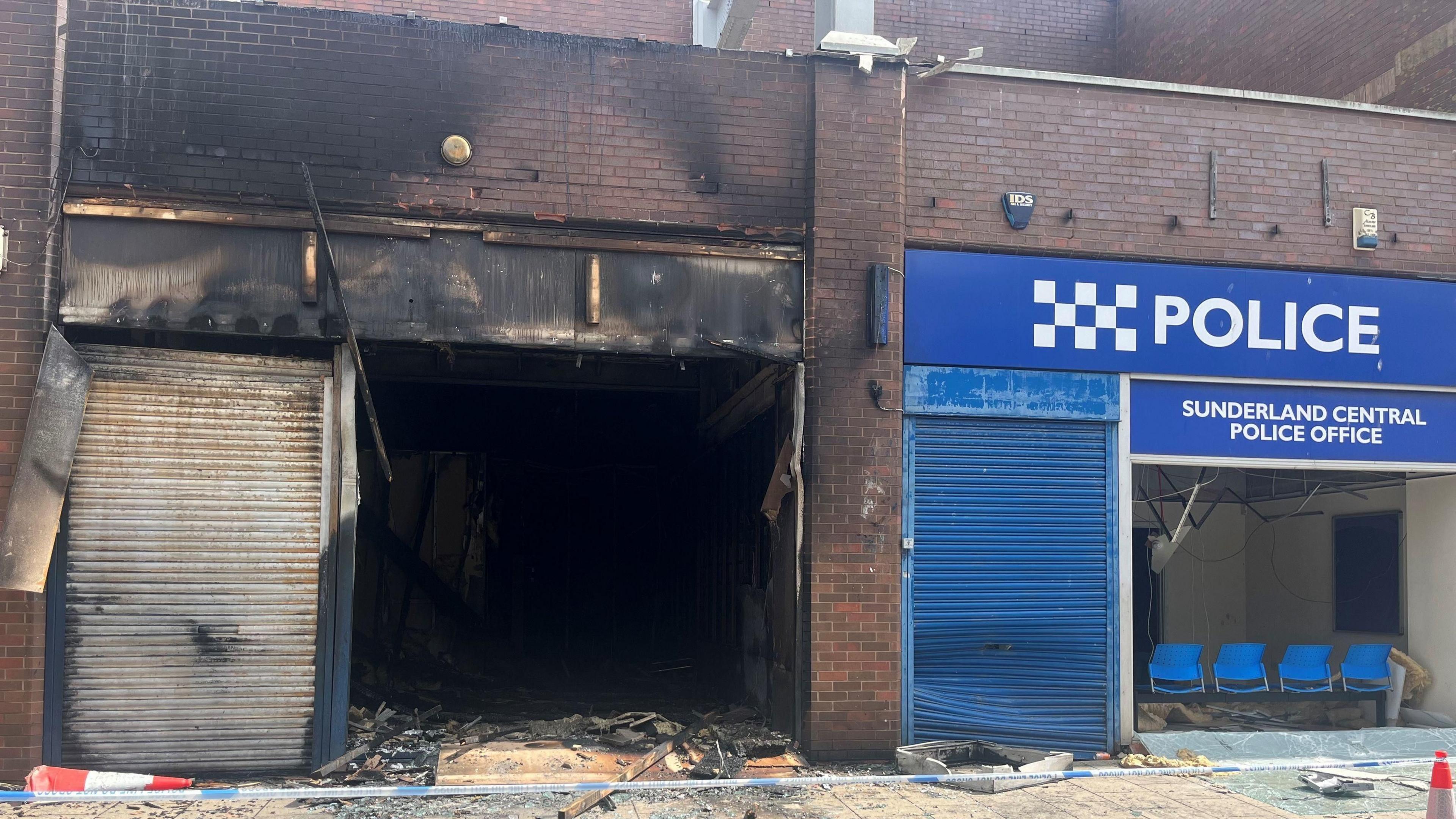 A burned out building next to blue-fronted building with signs reading "Police" and "Sunderland Central Police Office". There are burn marks on the bricks and shutters and police tape in front.