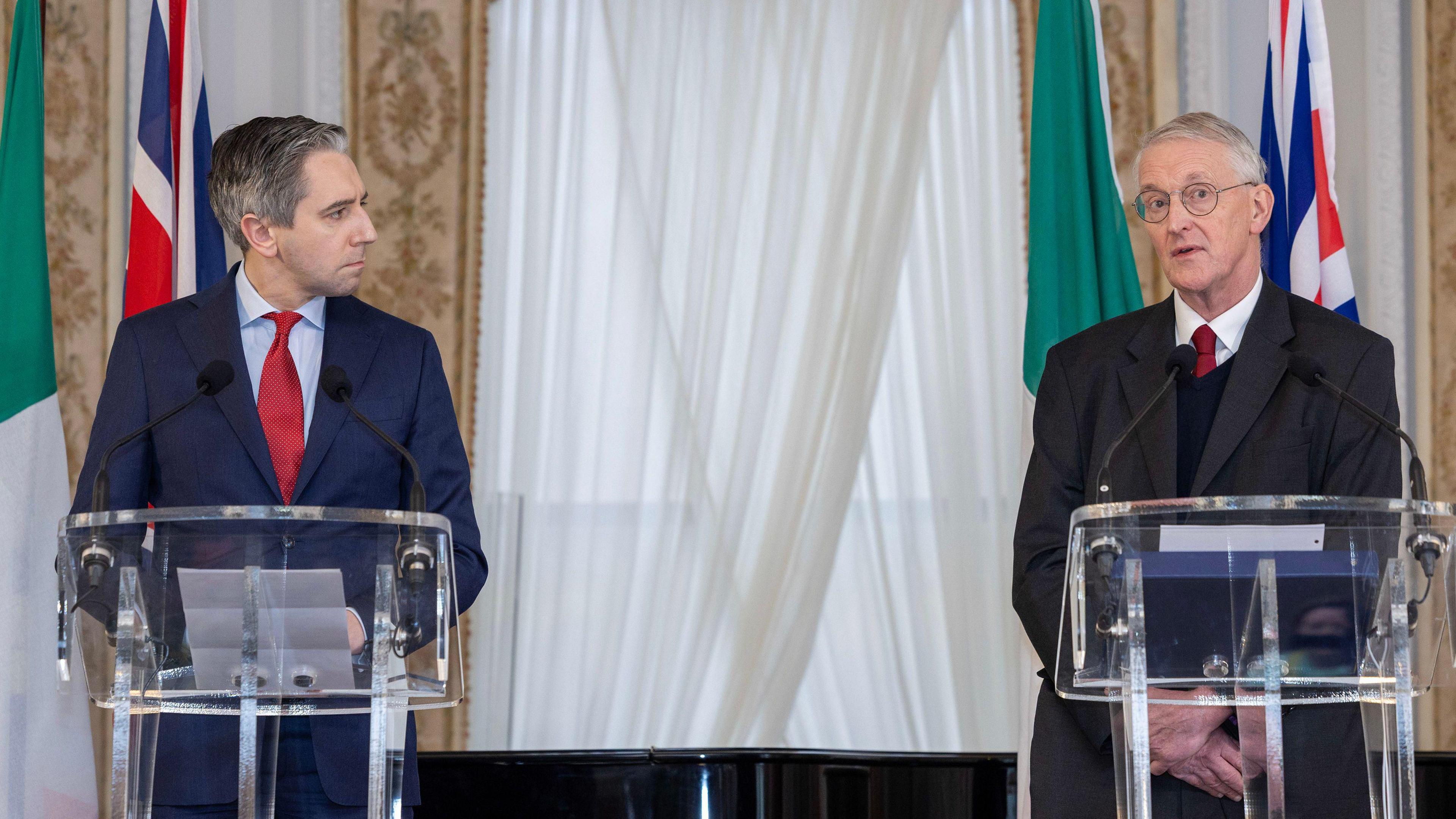 Harris and Benn are standing on a stage together. They are behind separate podiums with mics attached. The Irish and the union flags are behind them. Harris is wearing a navy suit with a red tie and Benn is wearing a black suit with a red tie. He wears glasses.