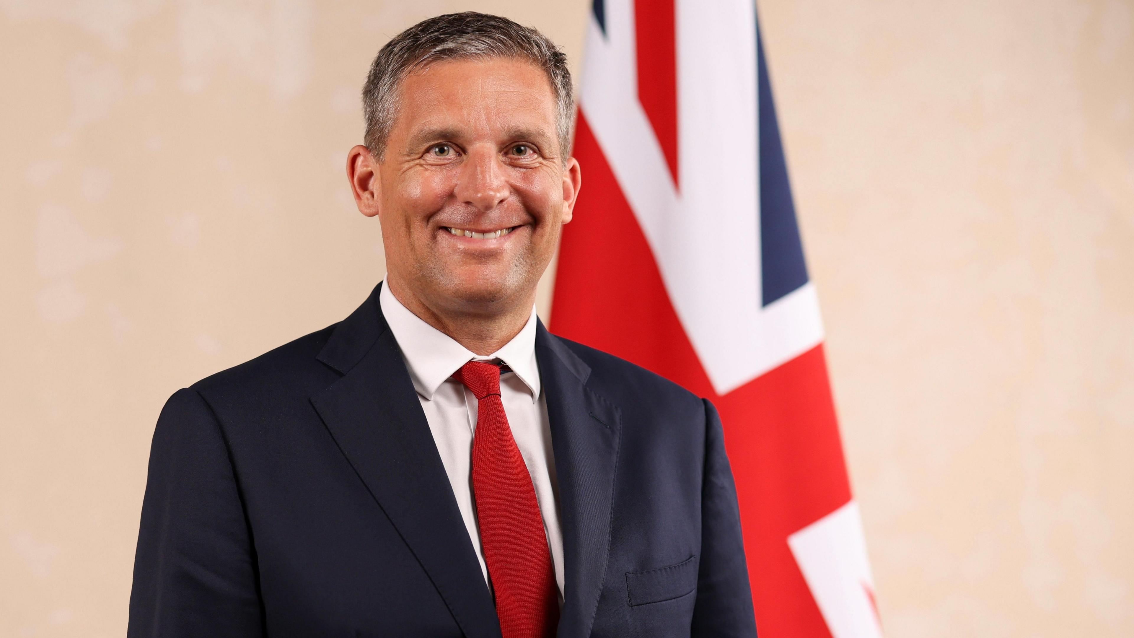 James Timpson is seen in an official photograph with a plain background and a Union Jack flag behind him.