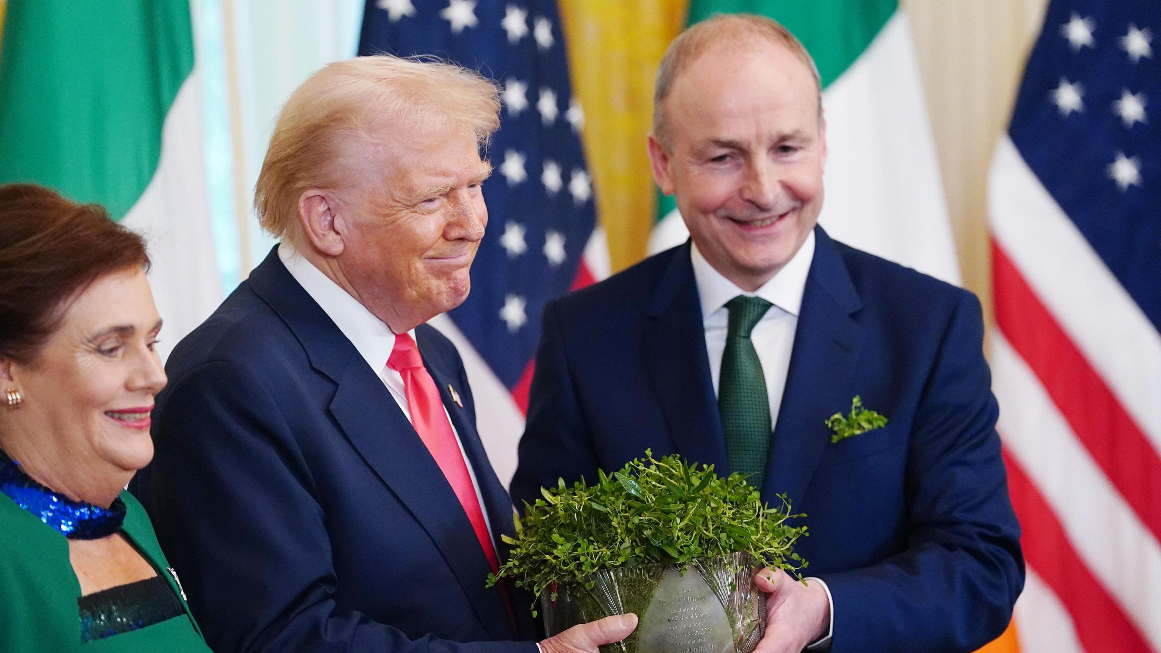 Micheál Martin hands US President Donald Trump a crystal bowl of shamrocks during a presentation at the White House. Behind the two leaders are multiple Irish tricolours and the US flag. To the left is Martin's wife.