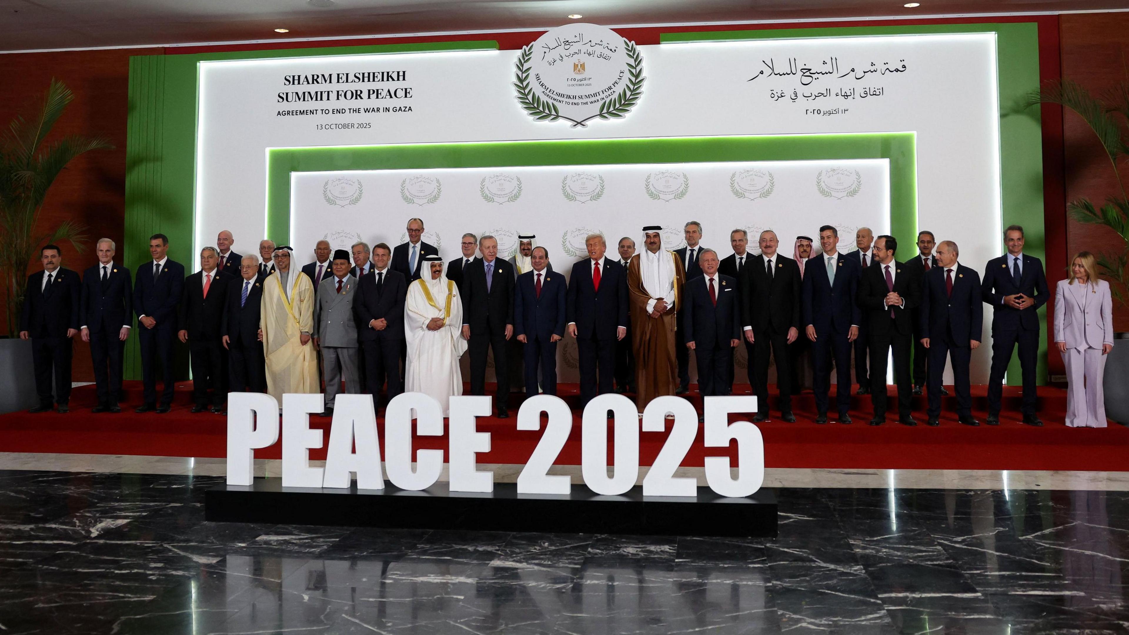 A row of people are stood on a red stage in front of white lettering that reads "PEACE 2025”.