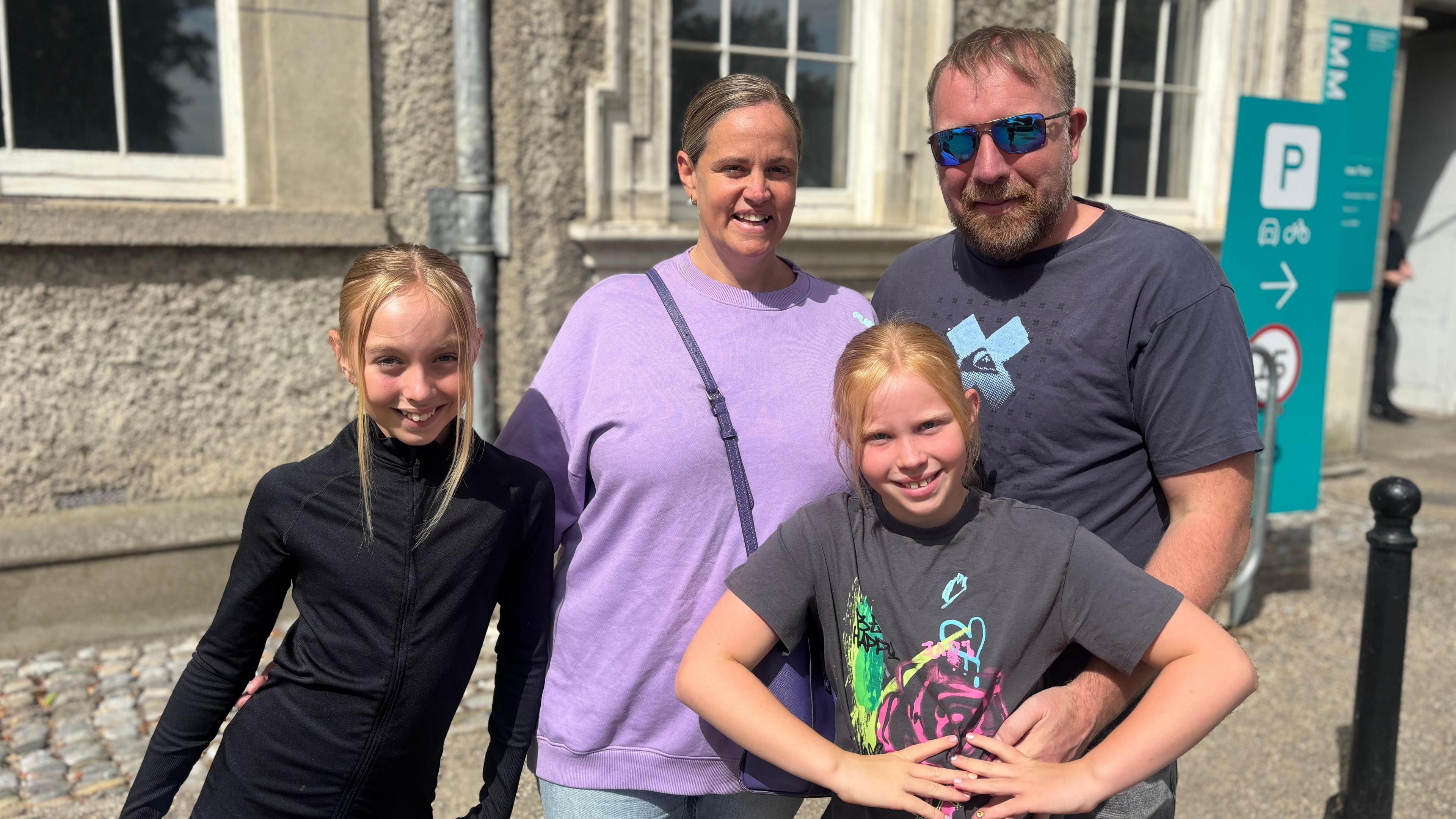 A family of four smile at the camera, the two girls have blonde hair, the one on the left is wearing a black top, the one on the right is wearing a grey top with blue yellow and pink design. Behind the woman is wearing a purple jumpers and man is wearing a navy top and sunglasses.