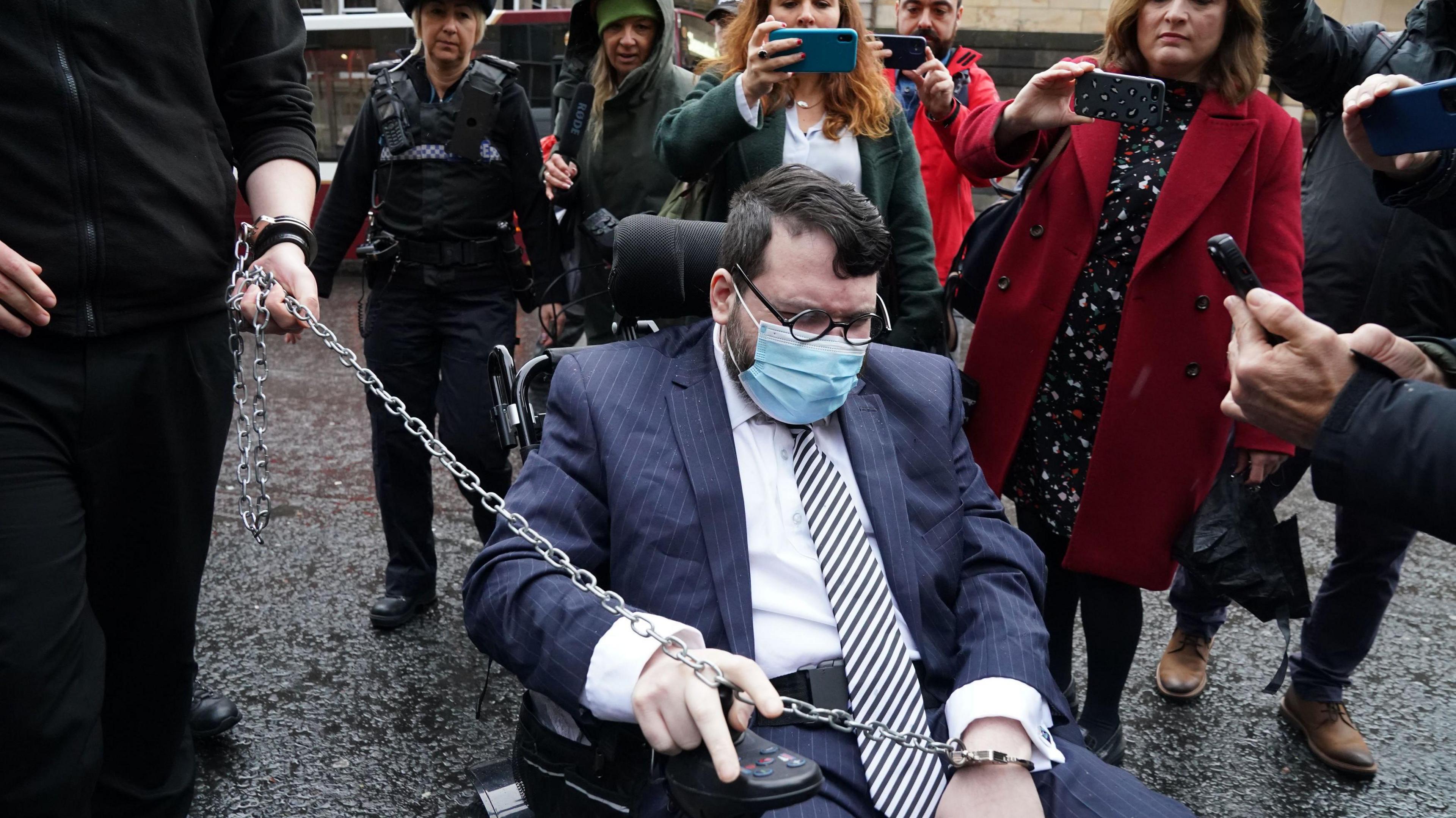 Nicholas Rossi outside court. He is in a wheelchair and is wearing a blue suit, white shirt, striped tie, glasses and an oxygen mask. People behind him are filming him on their phones and a long chain being held by a police officer links to his handcuffs.