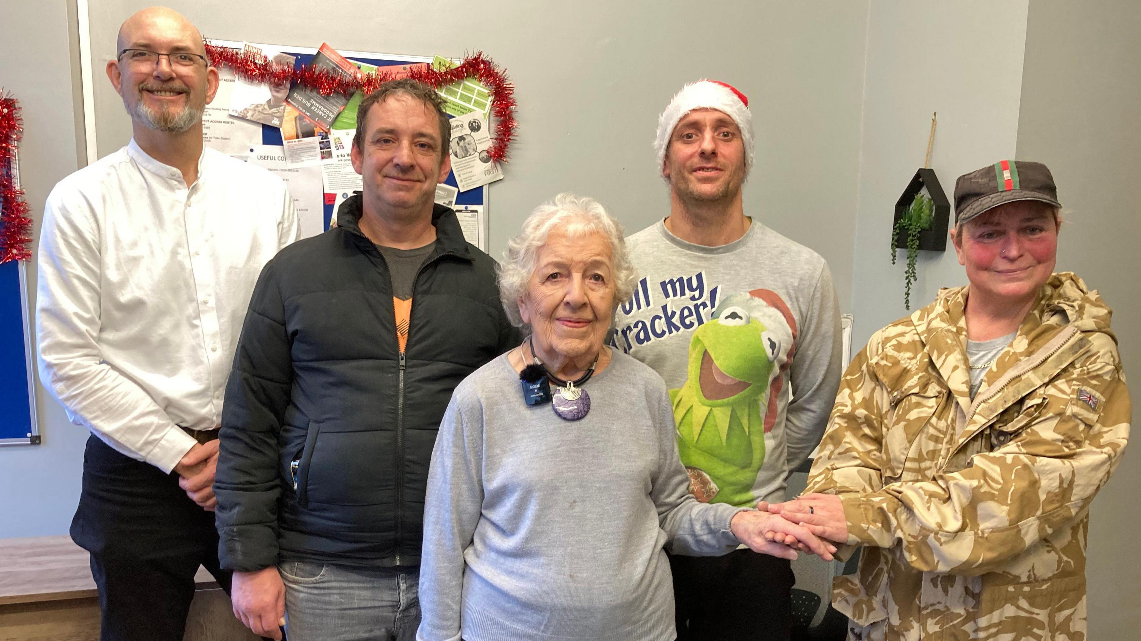 Three men and two women standing together. One man is wearing a Santa hat and Kermit the Frog on jumper. Two of the women are holding hands and smiling.