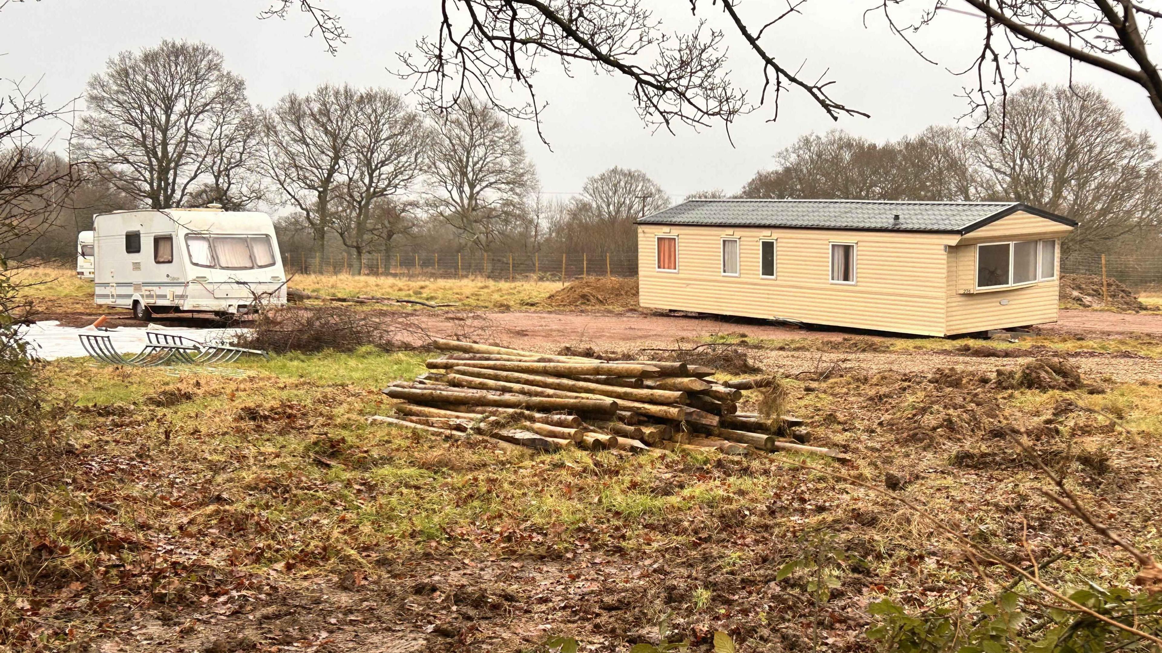 A static and a touring caravan on a field. A large pile of logs lies in the middle of the land.