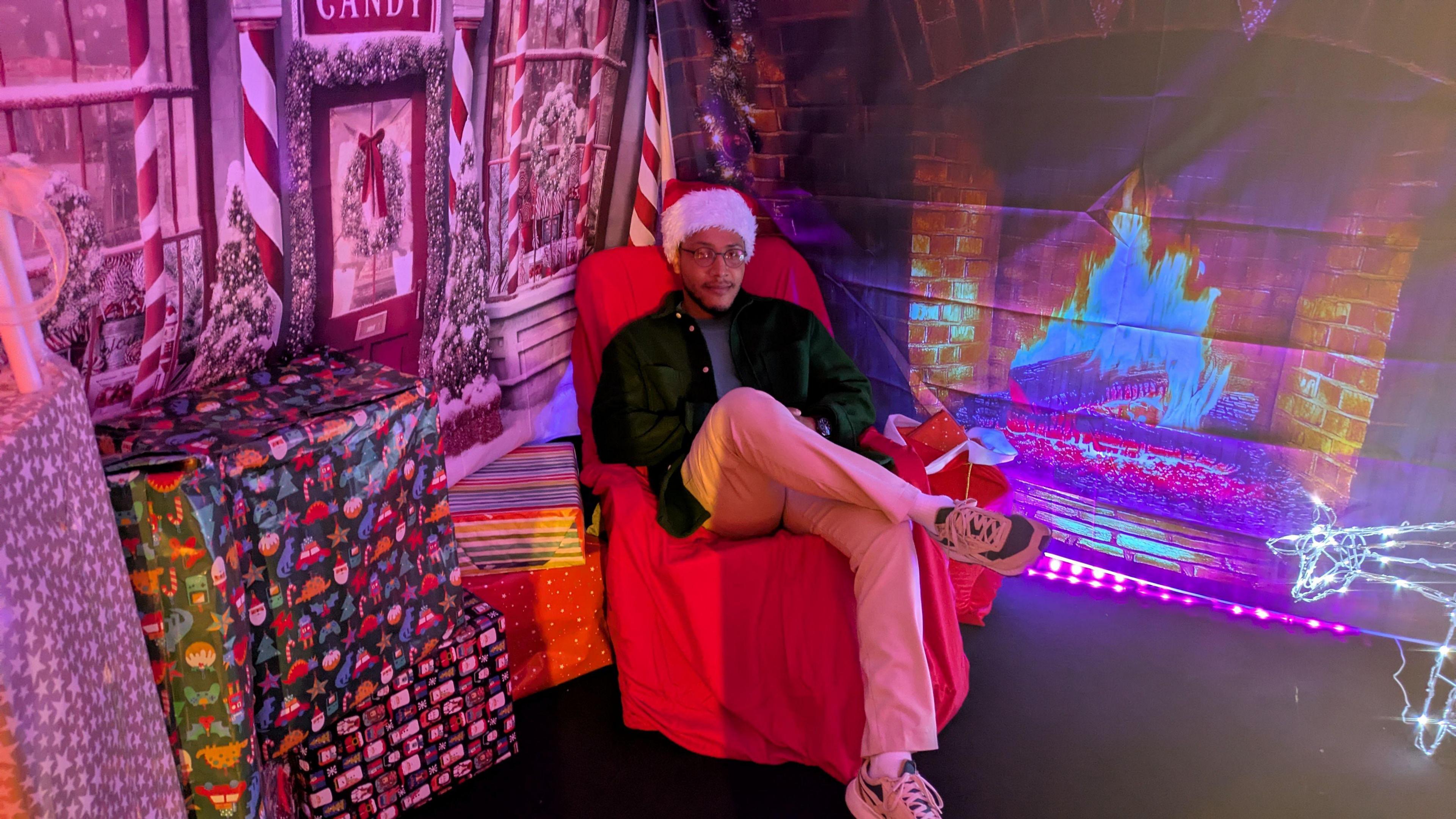 A man in a Santa hat sits in a red chair in a grotto, with sheeting up showing a burning fireplace and a candy store. Wrapped presents are stacked up in the grotto.