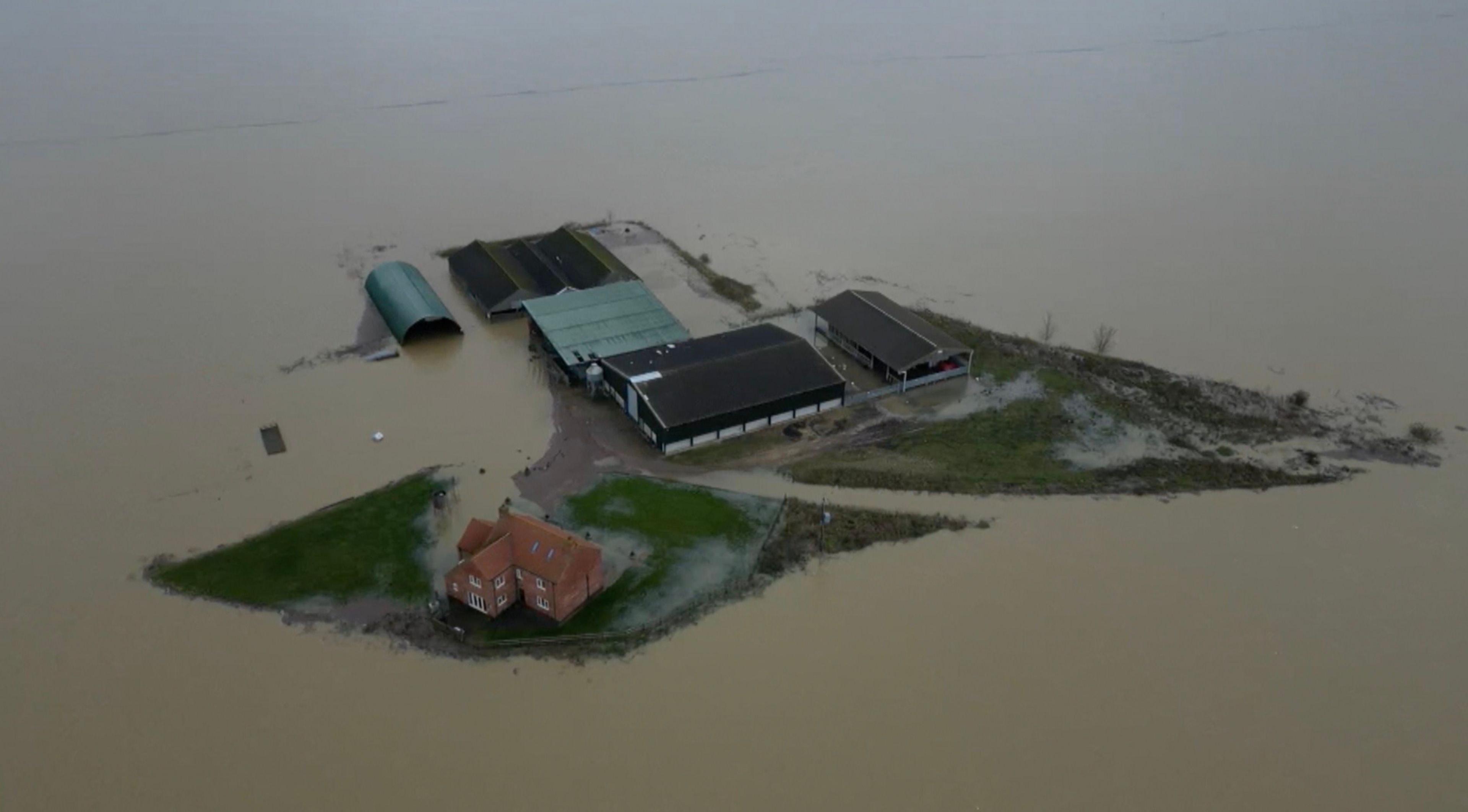 An aerial image of floodwaters surrounding a red brick farmhouse and barns, the water is brown and gives the impression of the farmhouse as an island. 