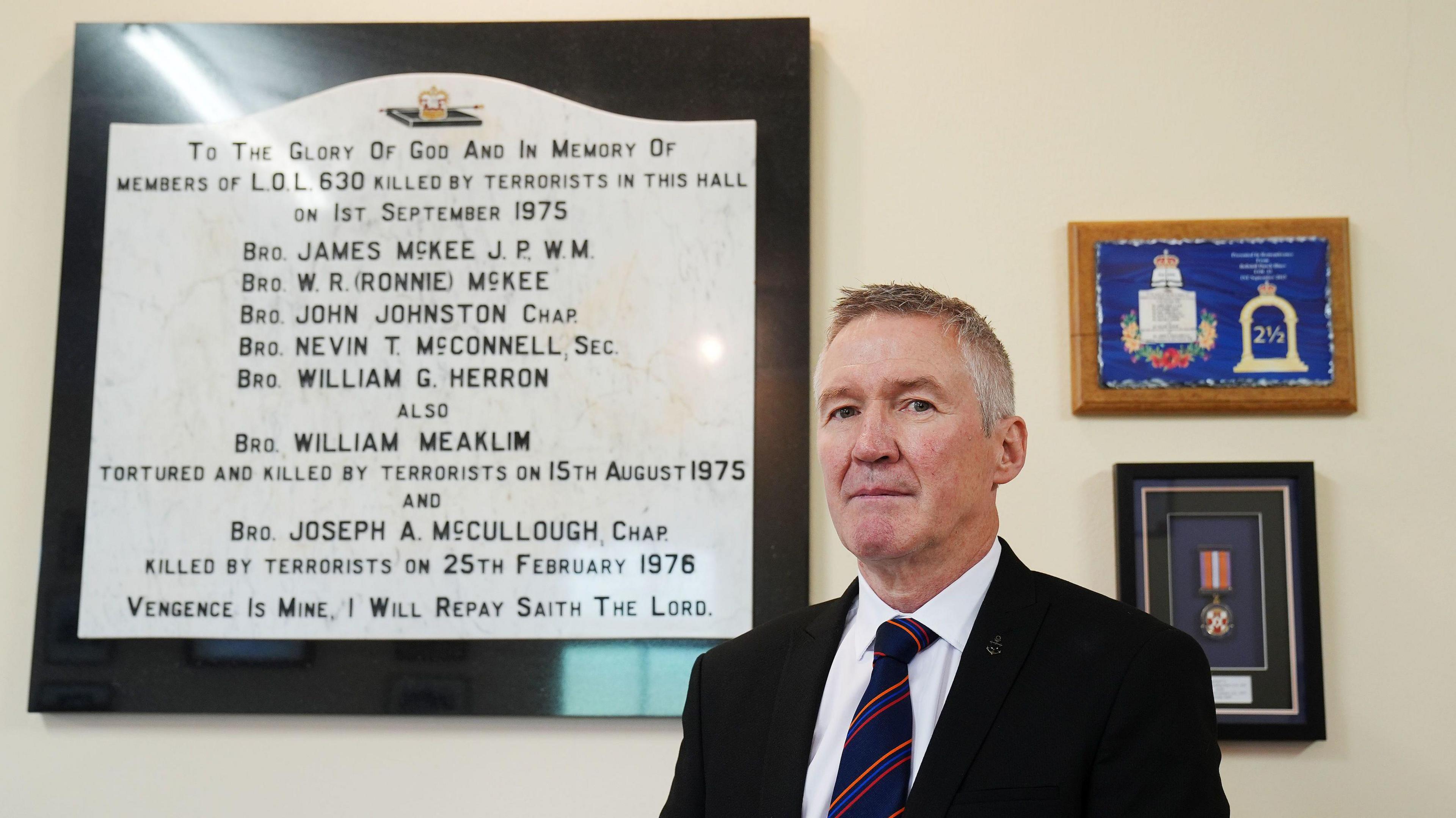 A man with short grey hair, wearing a white shirt, black suit and navy tie with red stripes, stands to the right of the image. There is a large memorial on the wall behind him with names of victims of the Tullyvallen attack written on it. 