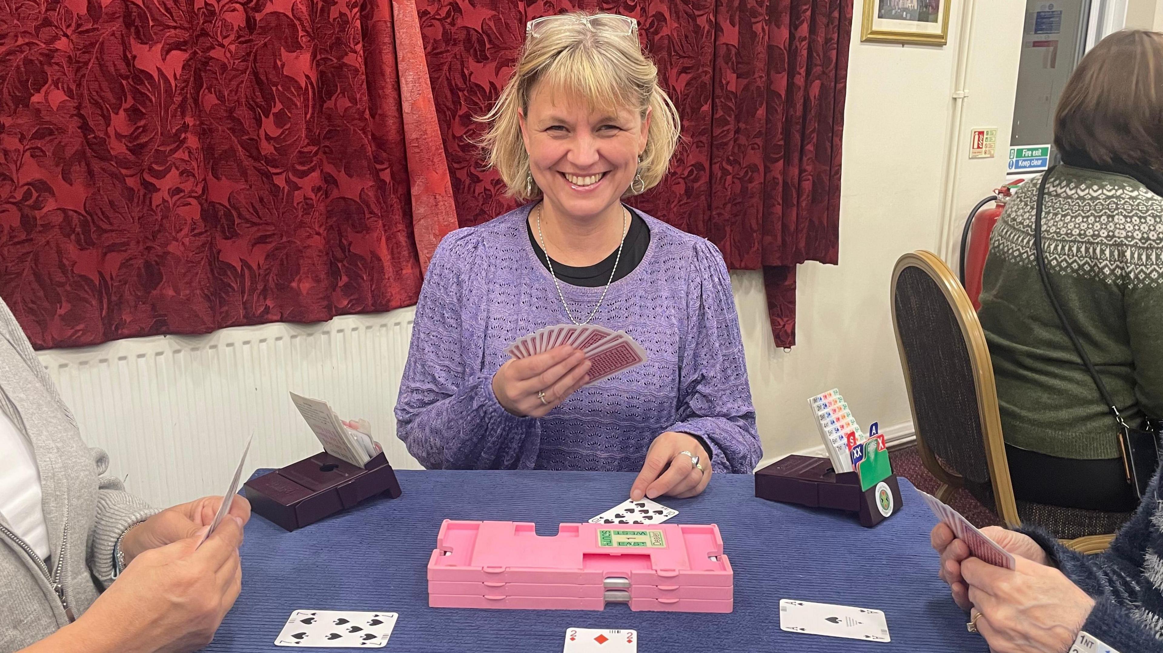 Abbey Smith sitting at a table playing bridge. A pink plastic box is in front of her, with a blue covering on her table. She is holding cards, with other cards in front of her. There are red patterned drawn curtains. She is wearing a purple top with a black top underneath. She has glasses on her head and has chin-length hair. She is smiling. There are two people at the table with her. A fourth person is sitting down, with their back to the camera, to the right