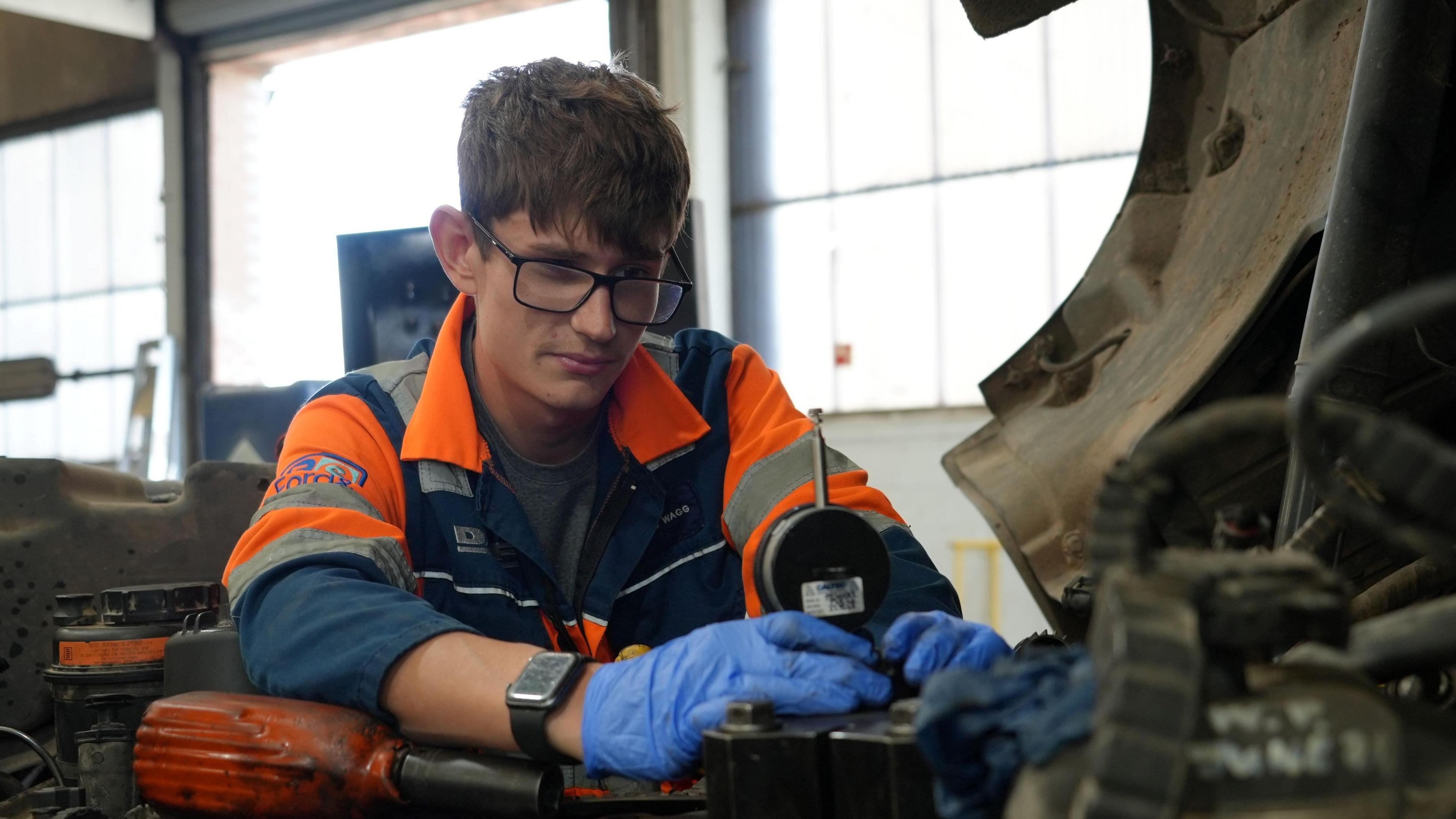 Charlie pictured hard at work in the garage in King's Lynn where he works. He has swapped his paddling kit for orange overalls and blue gloves as he fixes an HGV engine under the cab of a lorry.