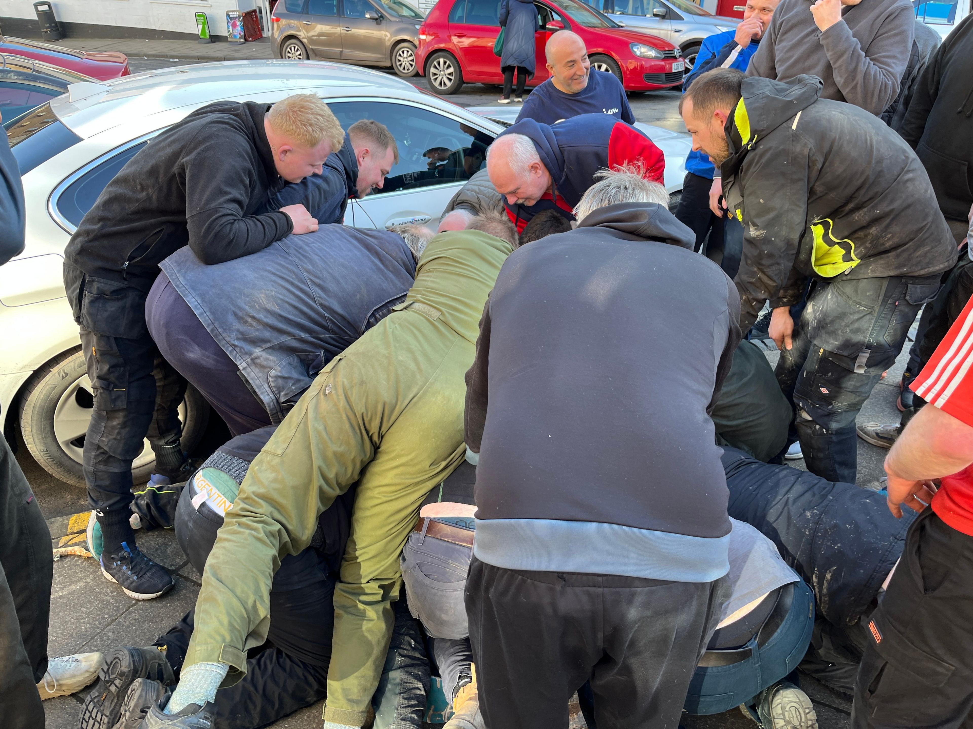 A group of men next to a car battle to get control of a small leather ball