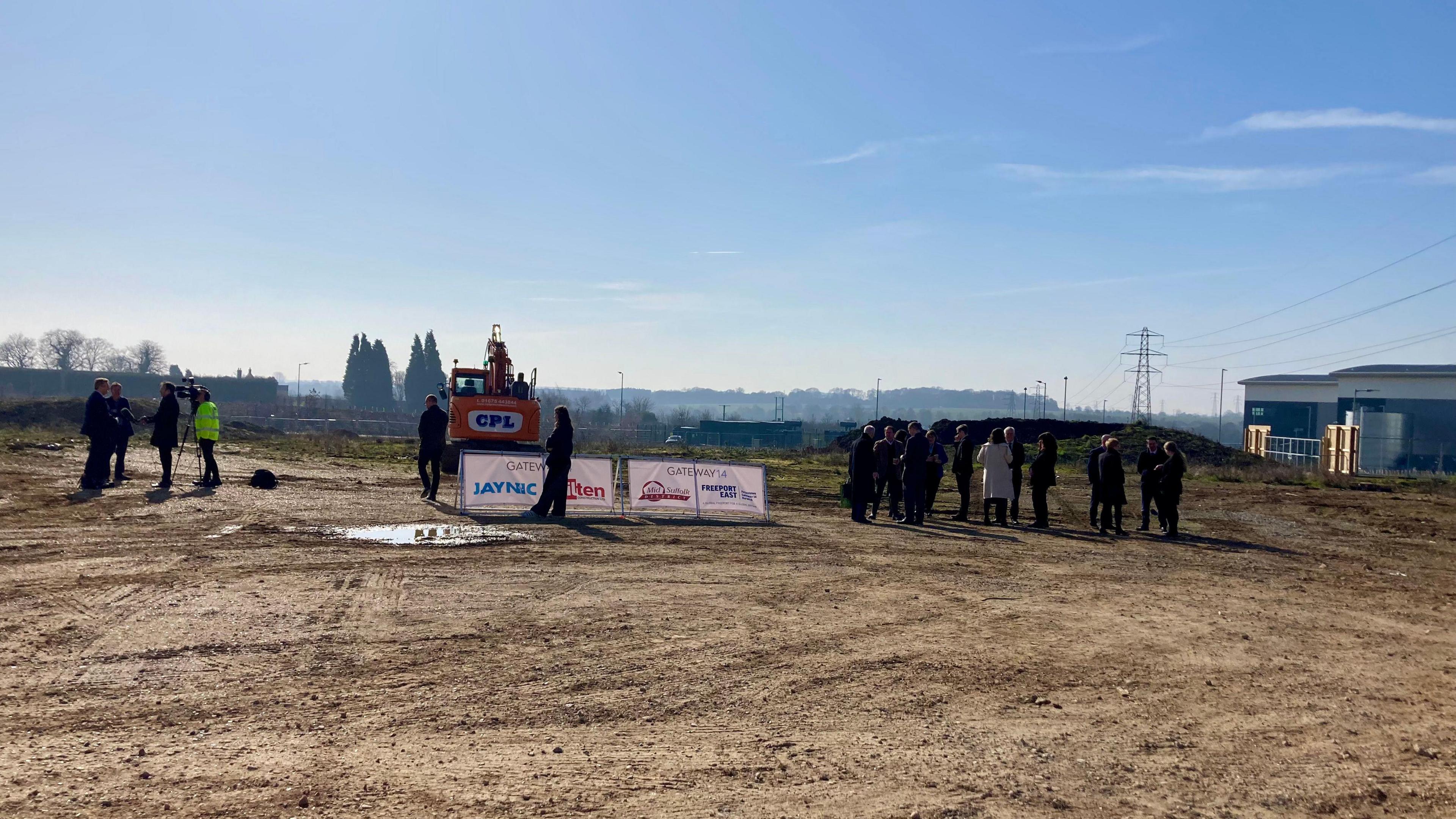 A large group of people stand to the right of the image. A digger rests in the middle behind two white signs. A camera crew interview some people to the left of the image. 