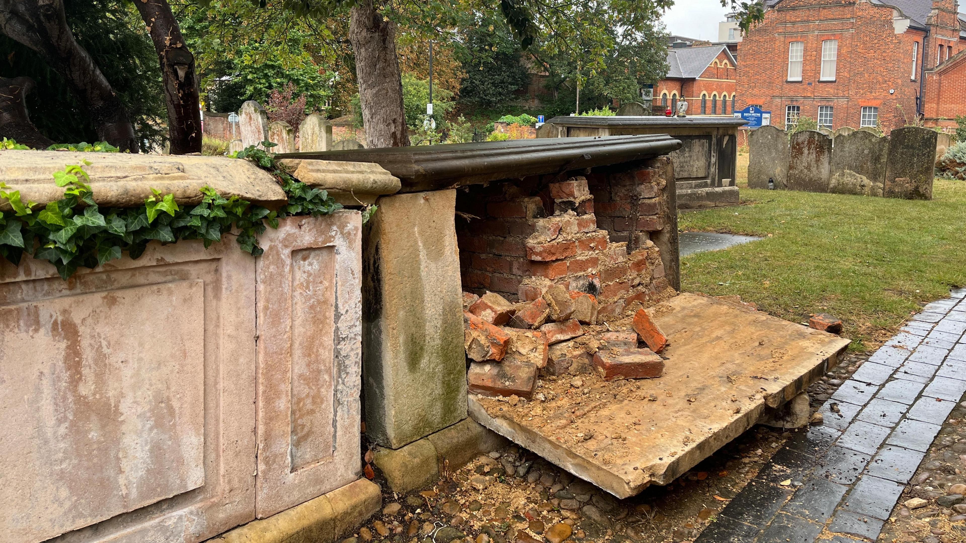 A tombstone in a graveyard which has one side of it torn open to reveal bricks inside. There is another tombstone next to it and gravestones in the background.
