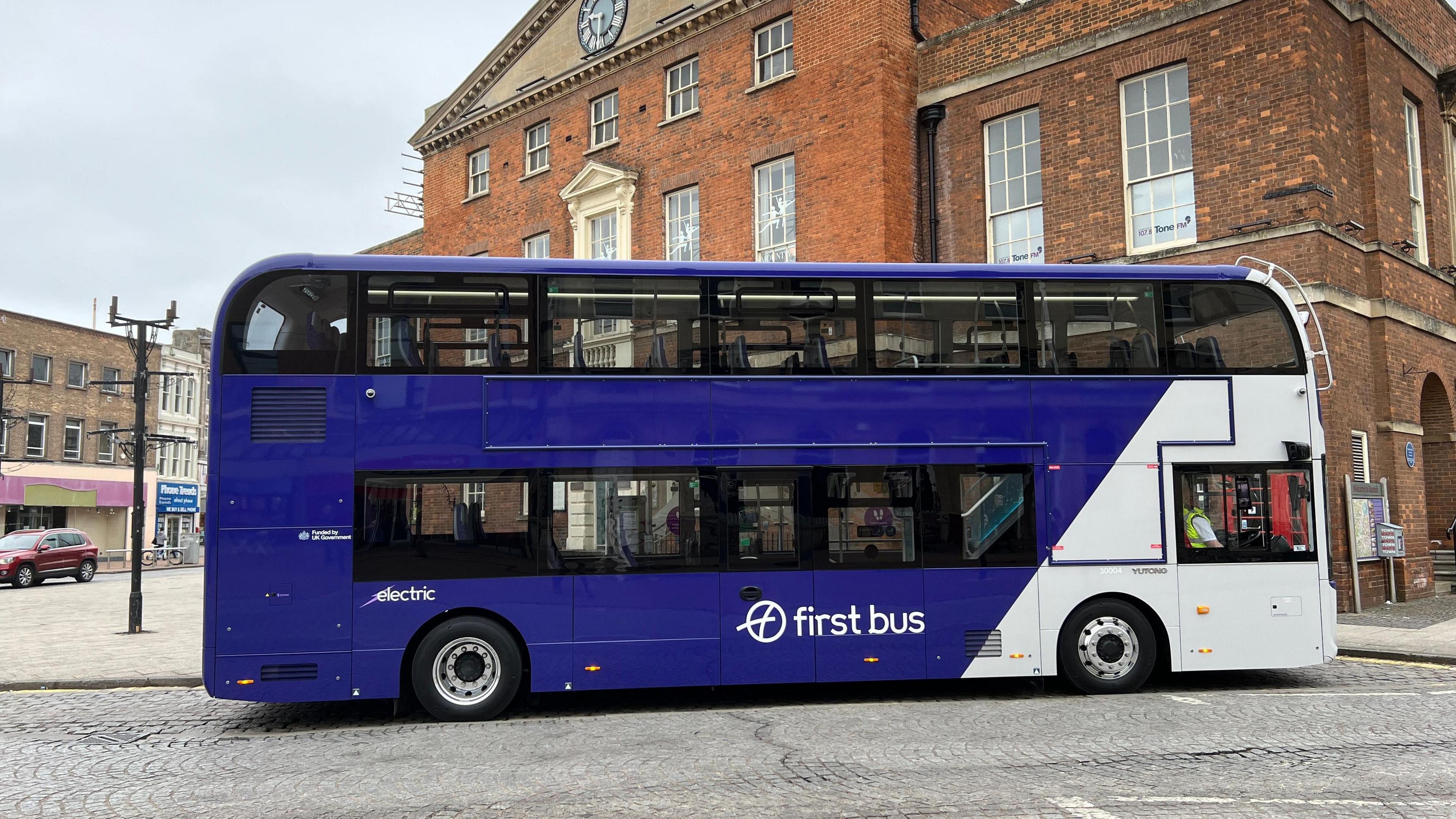 A first bus with blue and white branding. It is in the centre of Taunton outside an old victorian bricked building.
