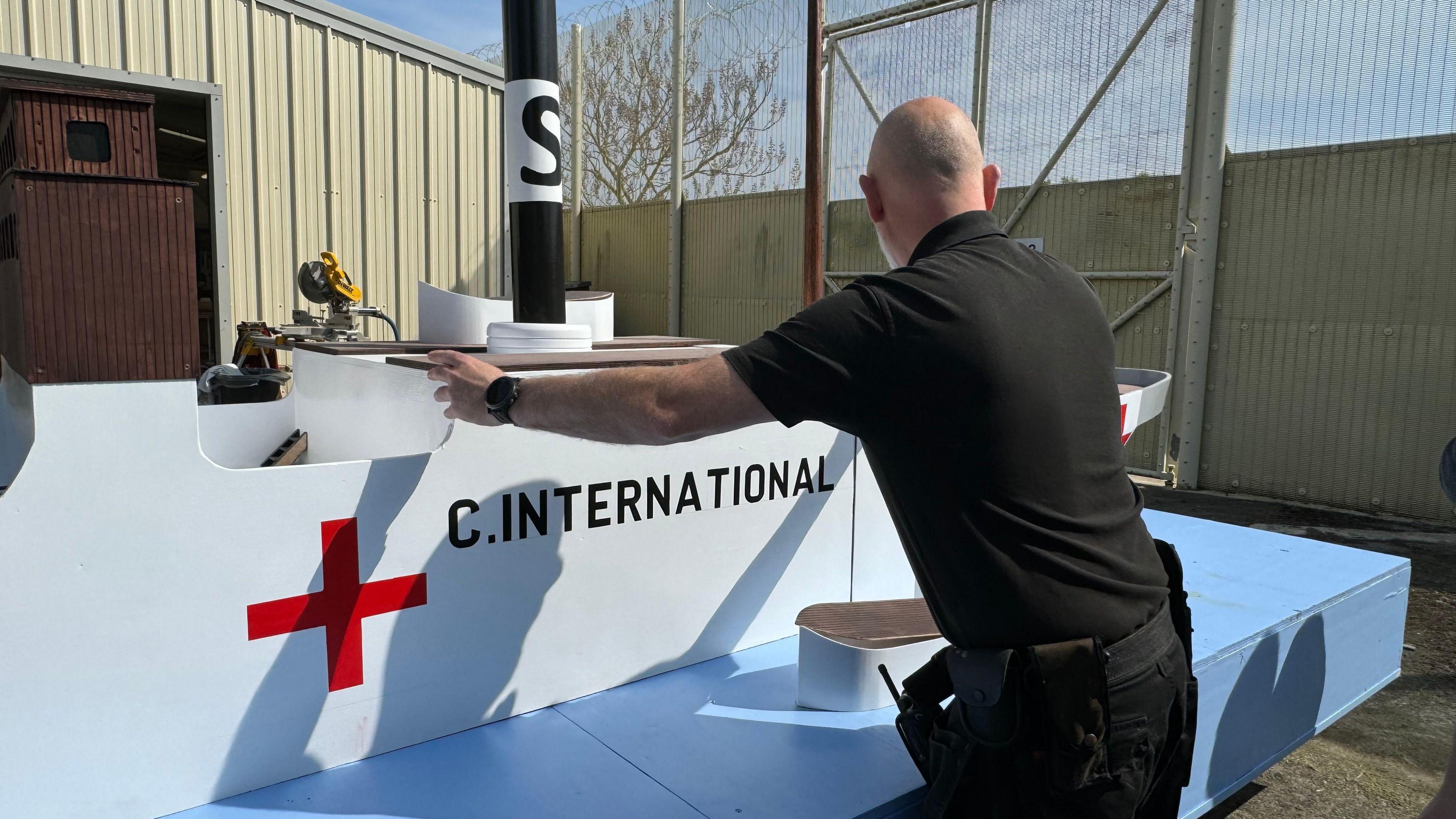 A man in a black top and trousers, facing away from the camera. He is touching the replica of the SS Vega. A model of a boat, the SS Vega. It is on a light blue board. The ship is white, with the words C.International on it and a red cross.
There is a prison gate behind them. 