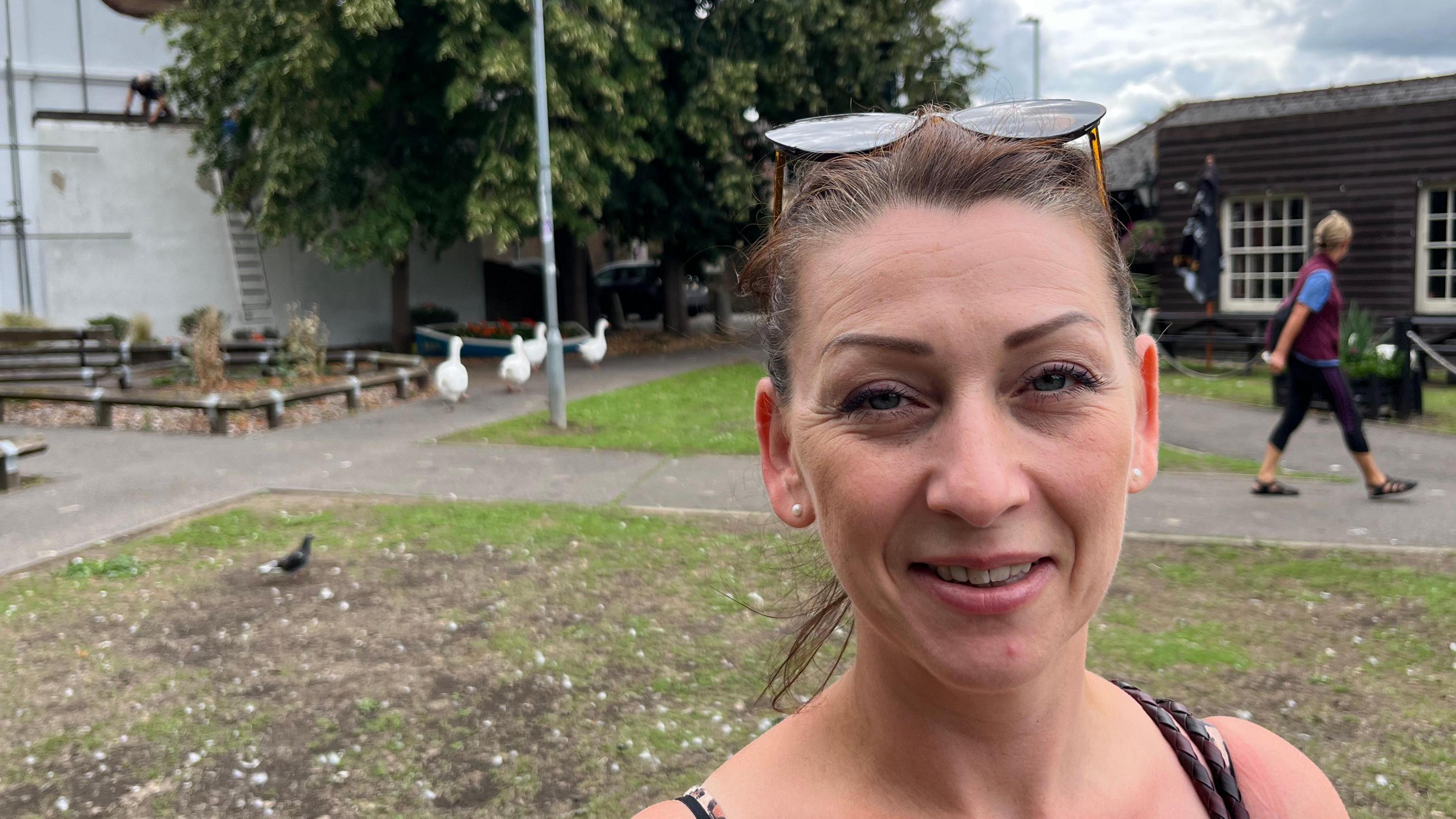 Kerry Howard, 43, has brown hair swept back tightly over her head, she has a pair of sunglasses perched on the top of her head and has gold stud earrings in both ears. She is wearing a strappy dress and her shoulders are exposed. A person is walking past on the right hand side and some geese are visible on the left.