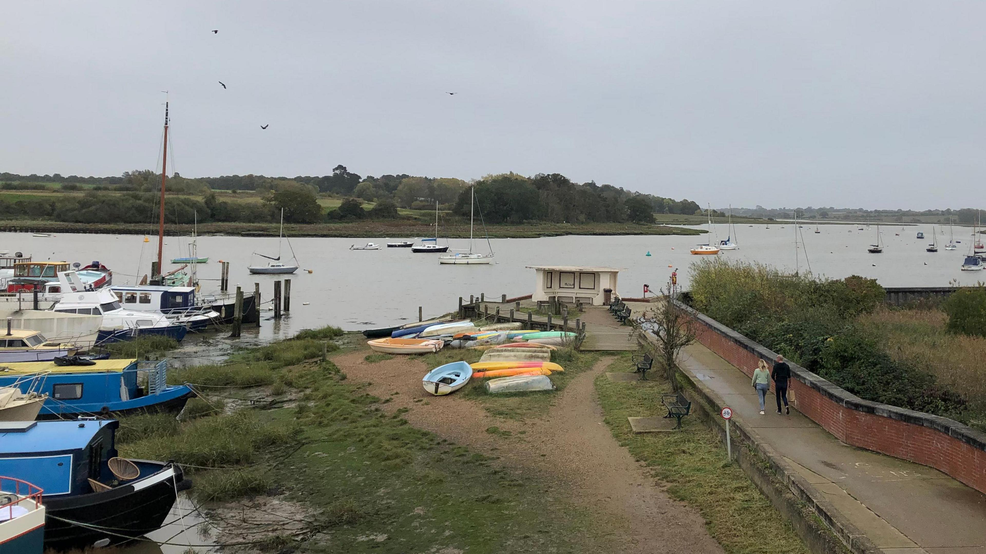 A selection of boats moor on the side of a river. In the distance boats are also moored on the river. On the right a man and woman are walking on a path.