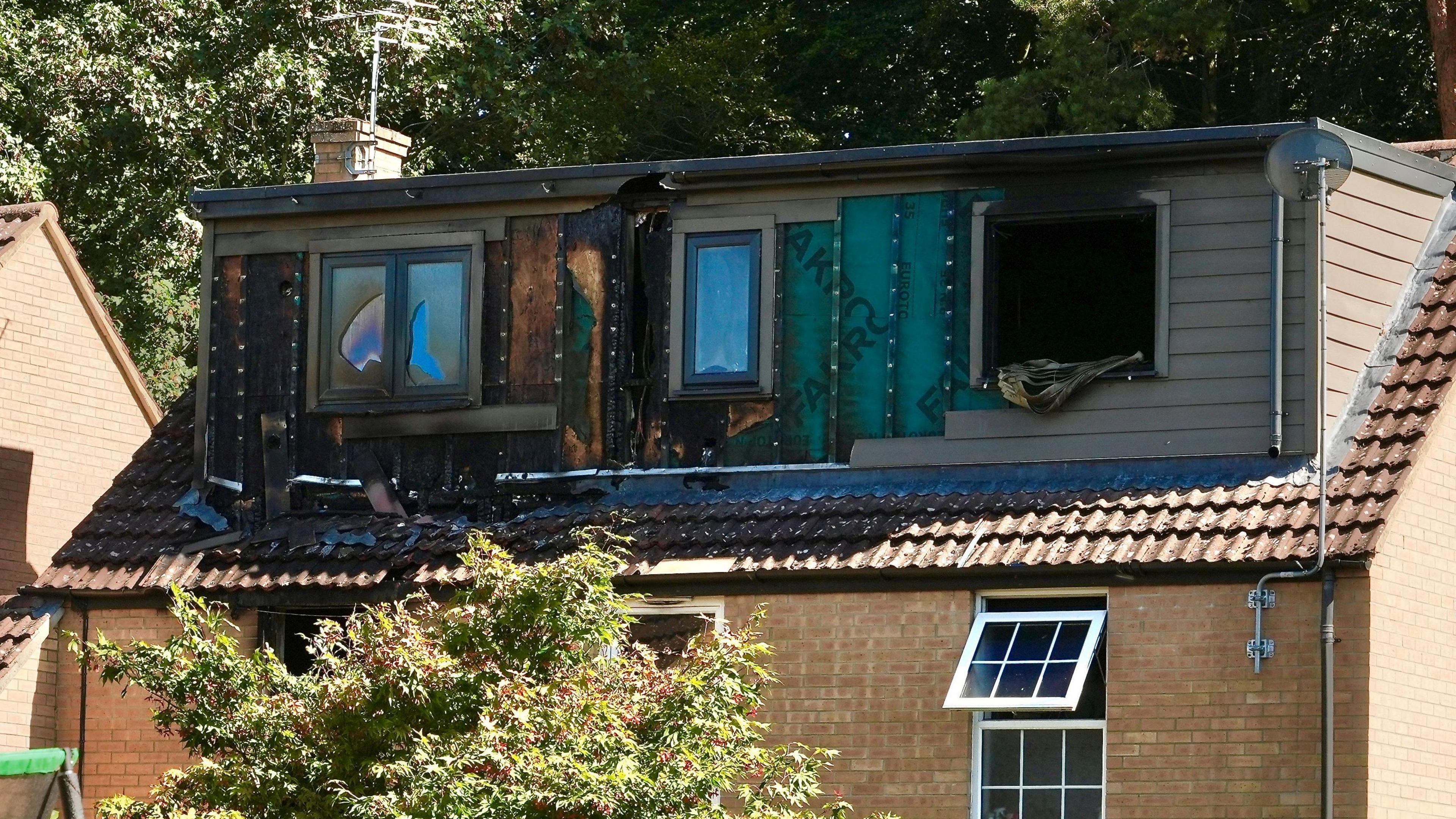 Three sets of fire-damaged windows can be seen as part of a loft extension on the roof of a sand coloured brick house, with trees seen in the background.