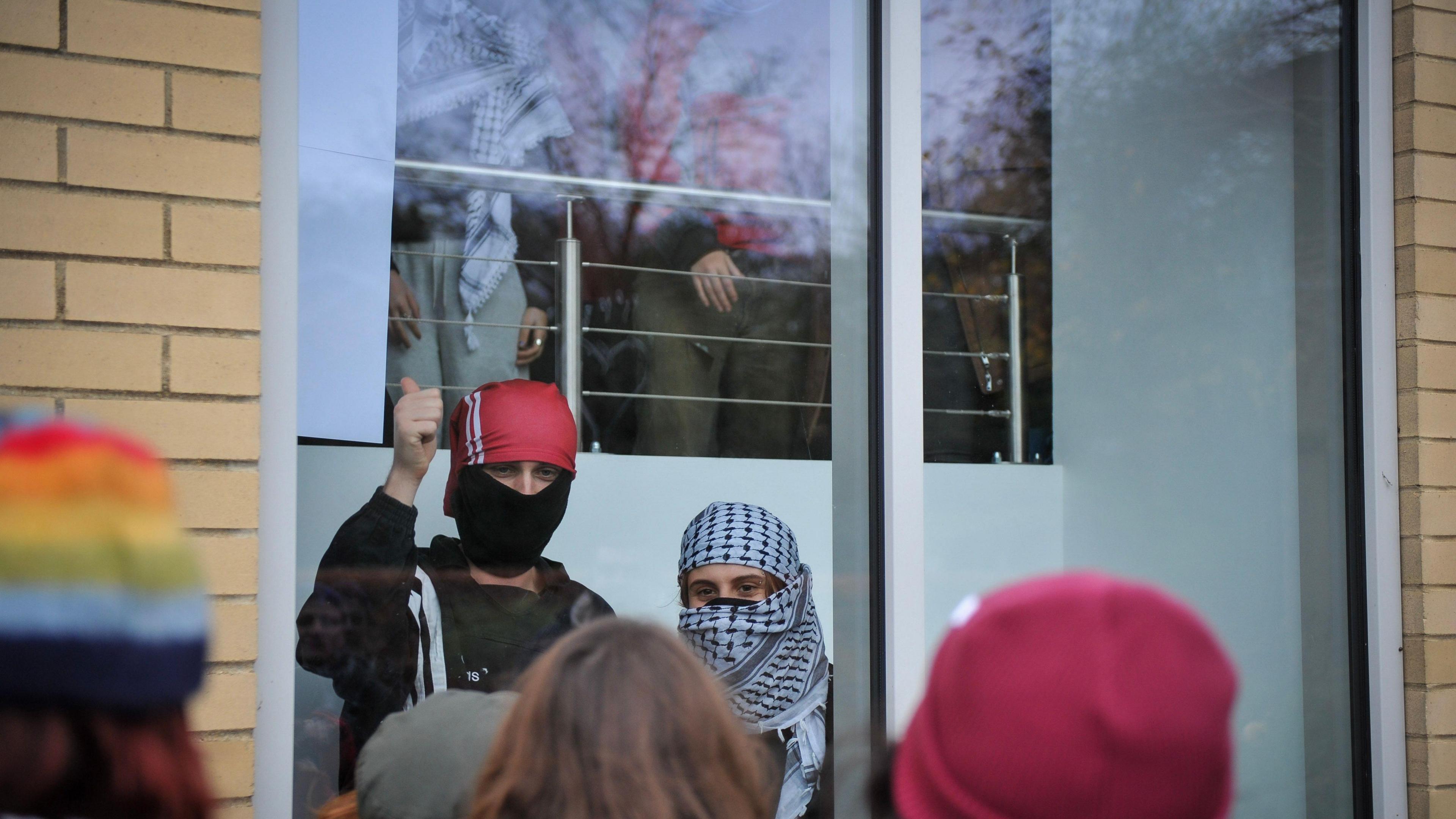 Protesters can be seen inside the building standing at windows. They have their faces covered and one of them is making a thumbs up sign. 