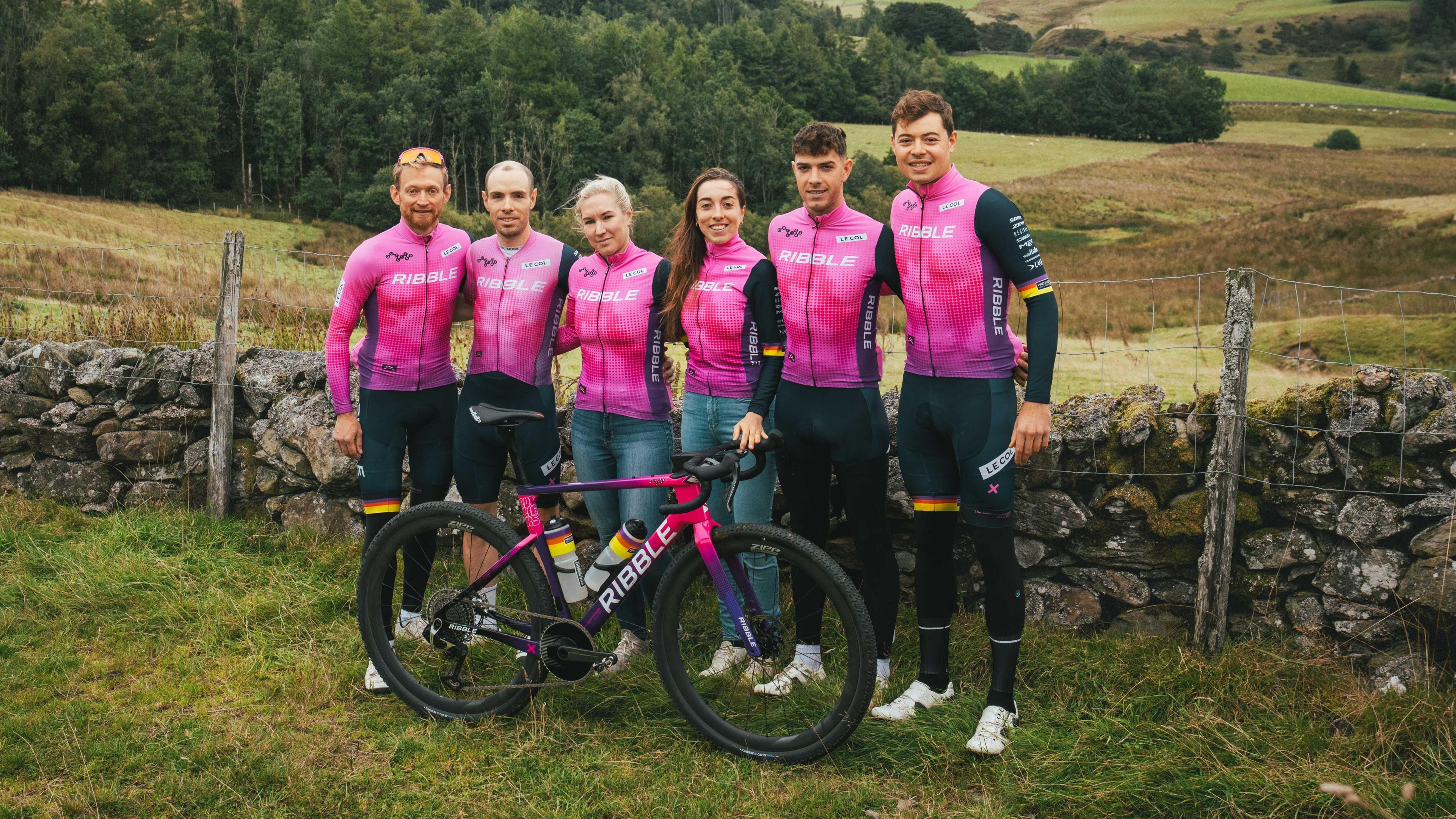 A group of six cyclists wearing matching pink and black Ribble team kits stand together in front of a stone wall and rolling countryside, with a pink Ribble-logoed bike placed in front of them.