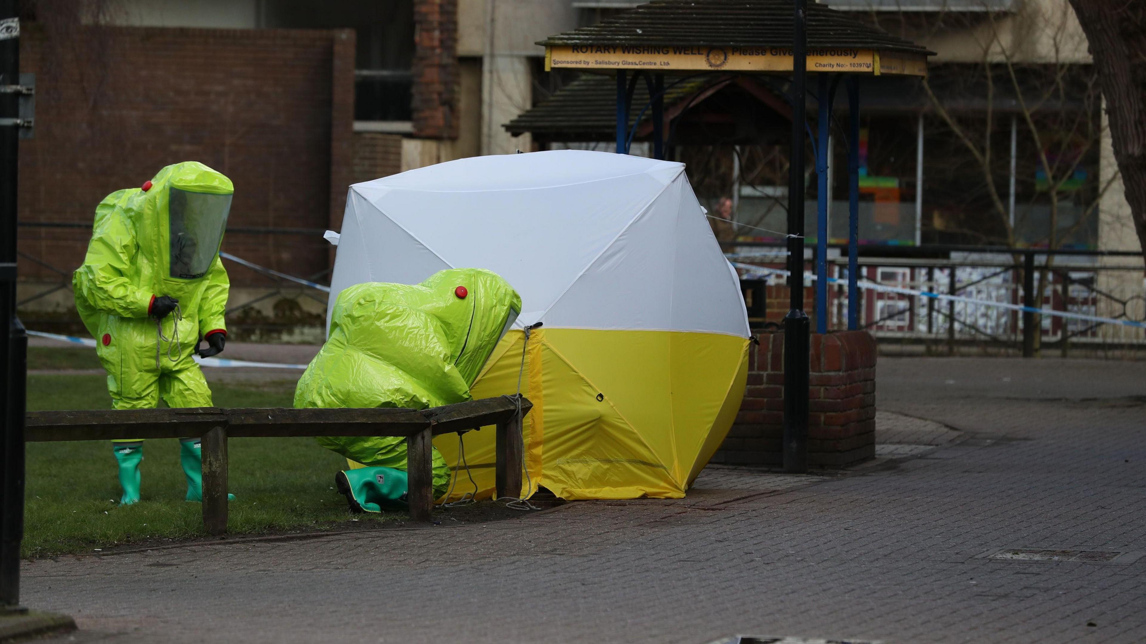 Two people in very large, bright green, hazmat suits, approach a yellow and white tent which sits over the bench where the Skripals were found. Police tape is visible in the area 