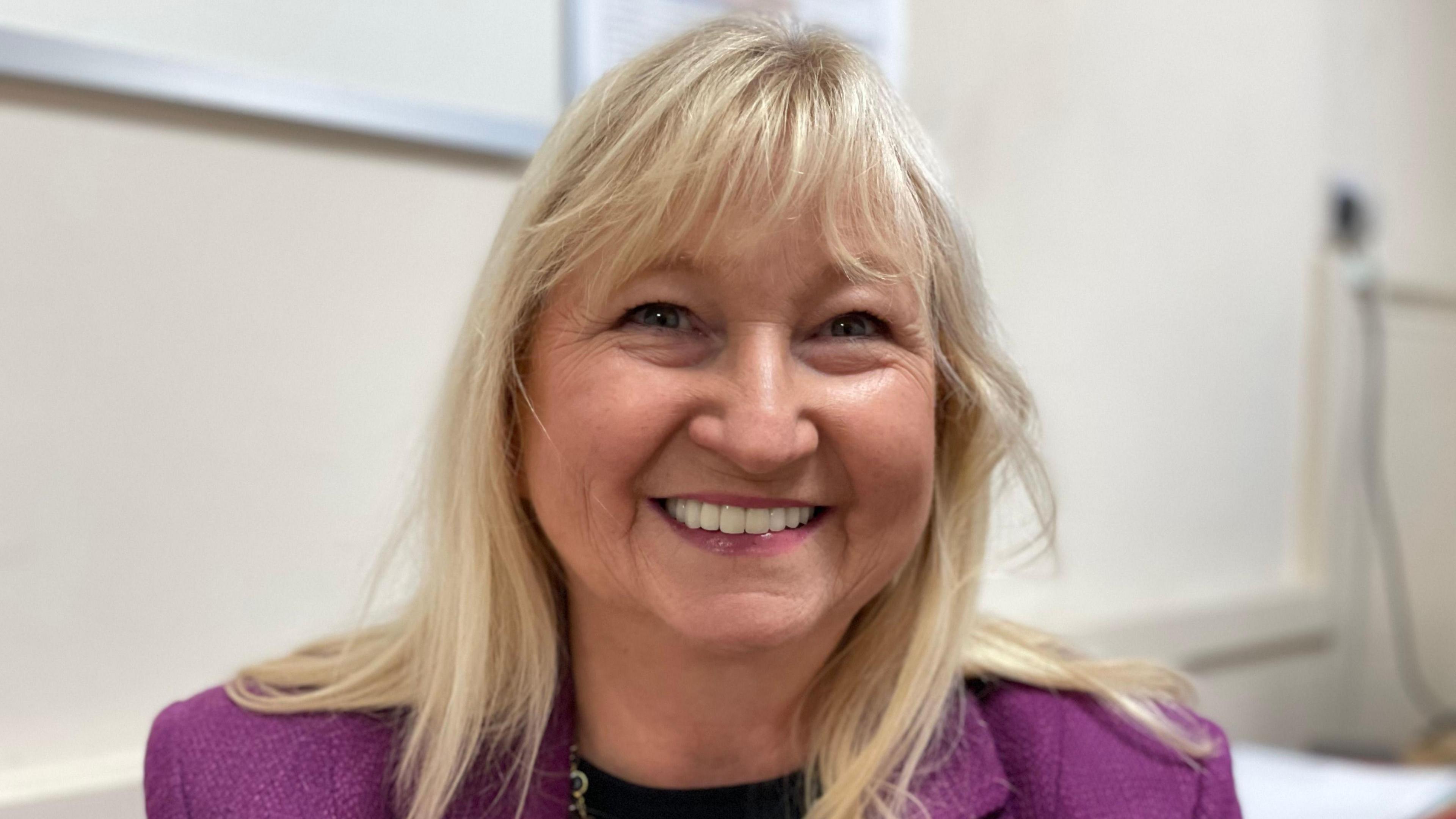 A head and shoulders shot of Council leader Anne Dorrian. She is wearing a black top and a purple blazer. She has blonde long hair and is smiling at the camera. 