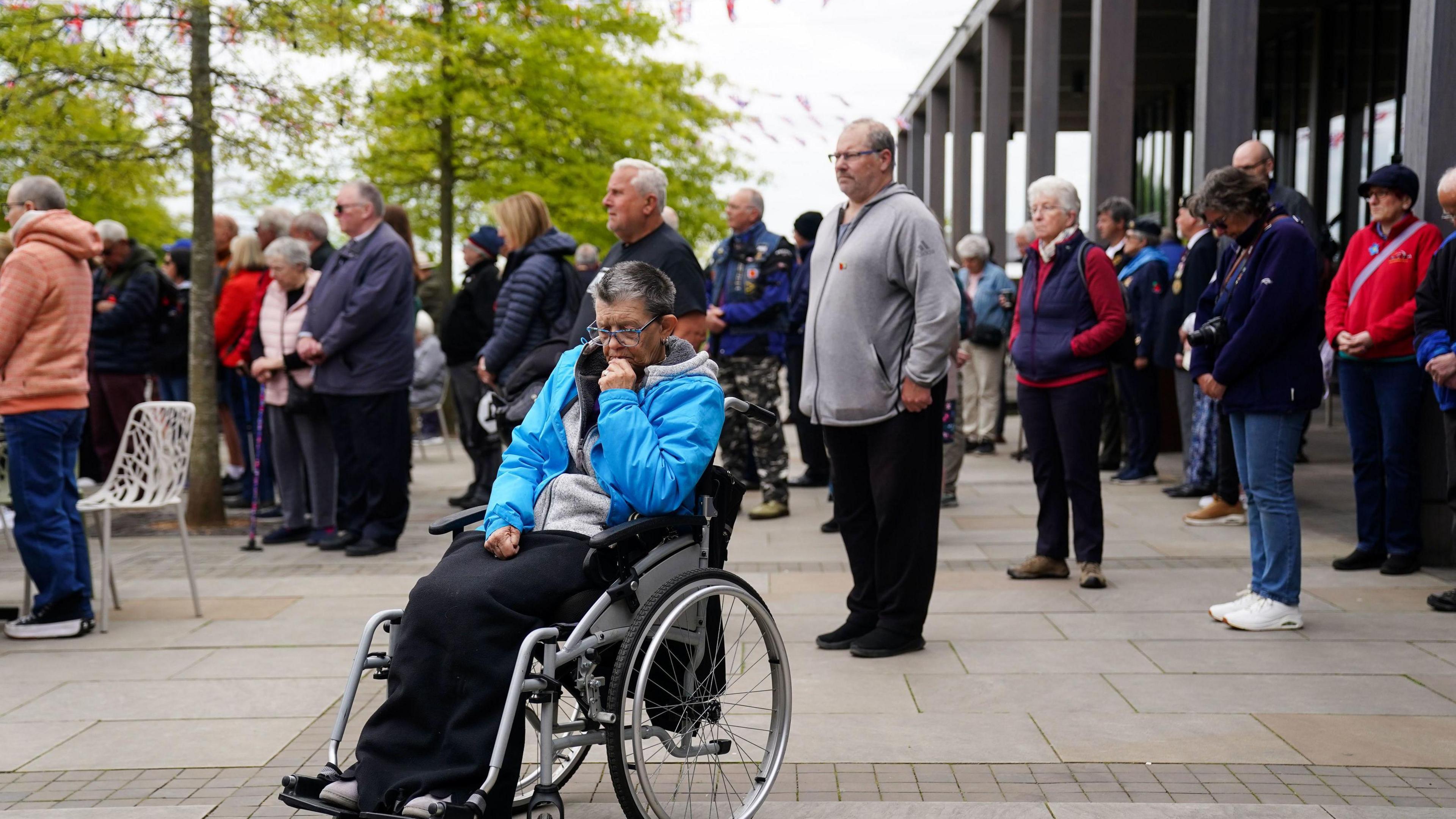 A woman in a wheelchair appears to be deep in thought as people stand silent to mark the two-minute silence.