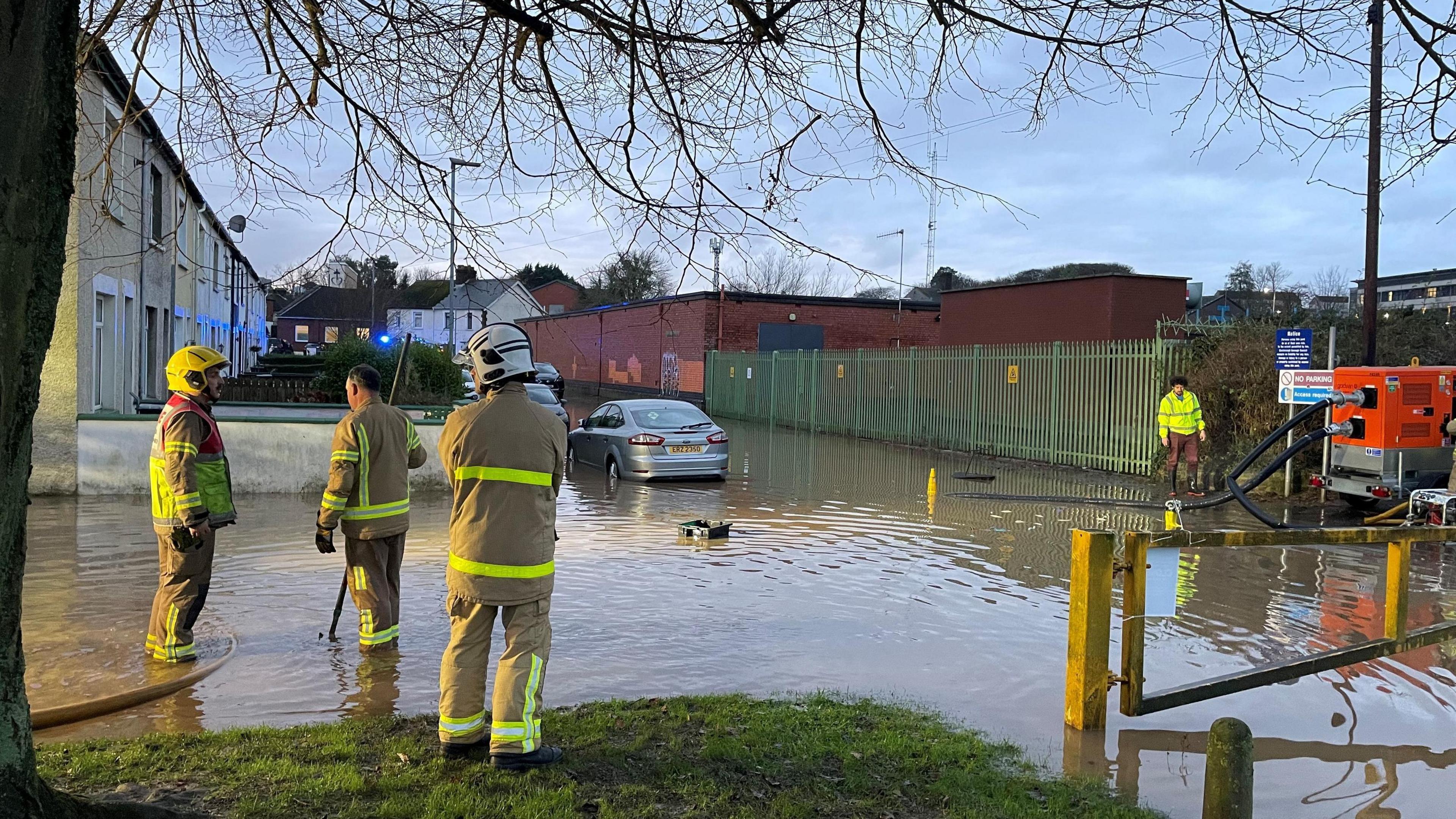 A flooded road with housing on the left and a red building and fencing on the right. There is a silver car on in the water and to the left there are three firefighters. 