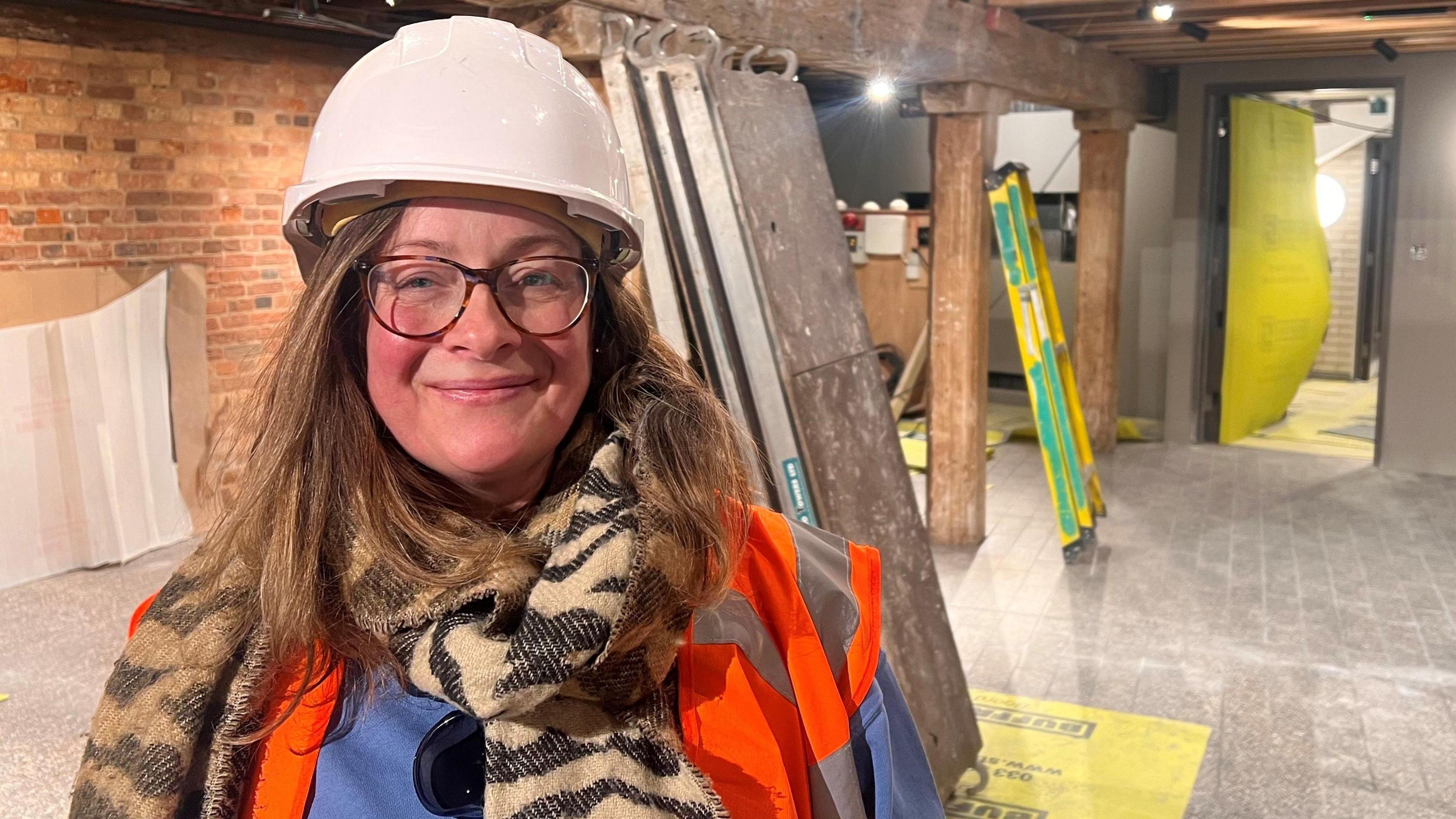 Alison Gudgeon wears a hard hat inside Poole Museum, which is stripped back to the brick walls with building materials visible behind her. She wears glasses and a large brown scarf and has long brown hair.