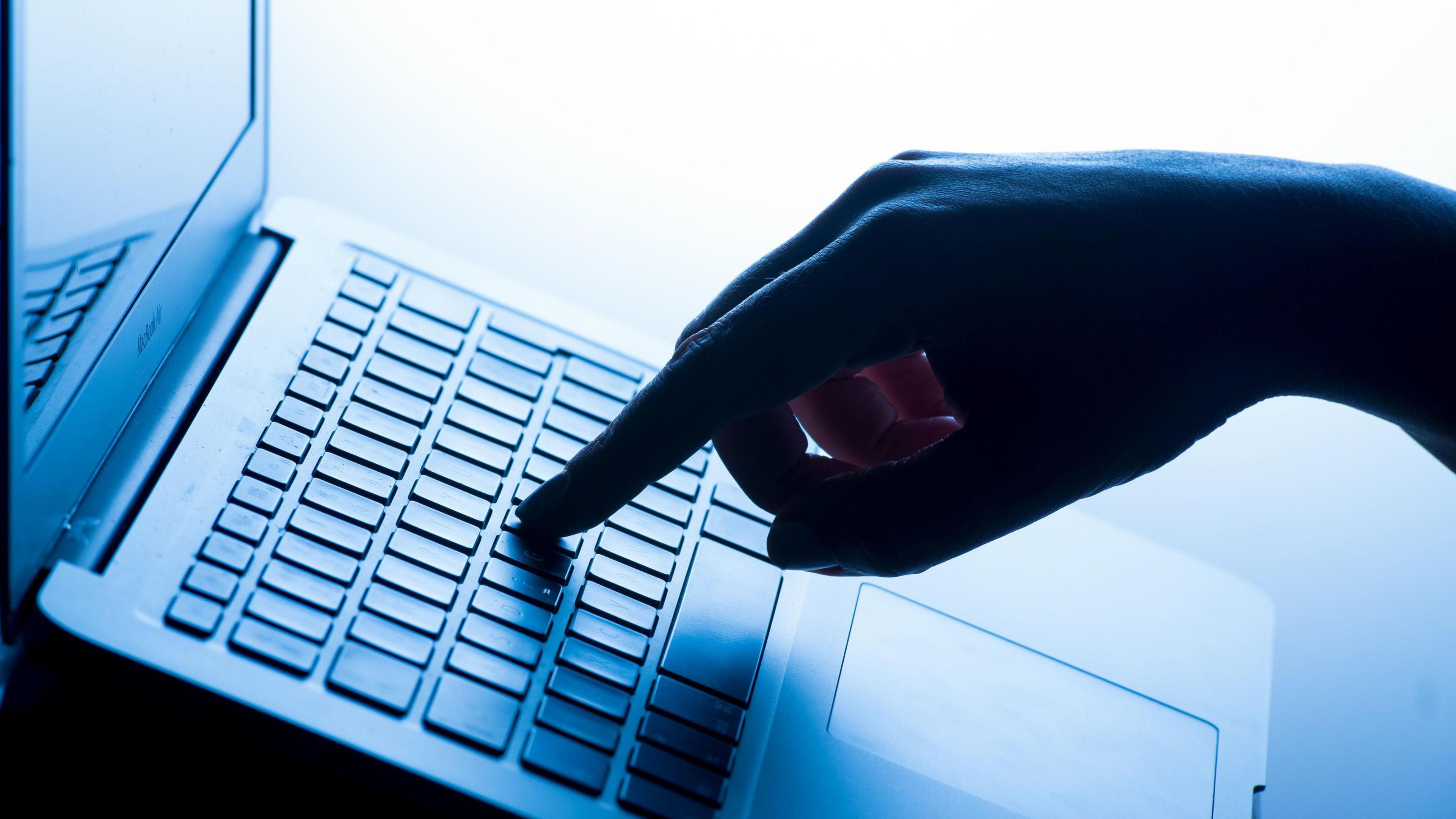 A silhouette of a hand typing on a laptop keyboard. There is a finger on one of the keys and the lighting is low and has a blue tone. The hand is on the right and the laptop is on the left.