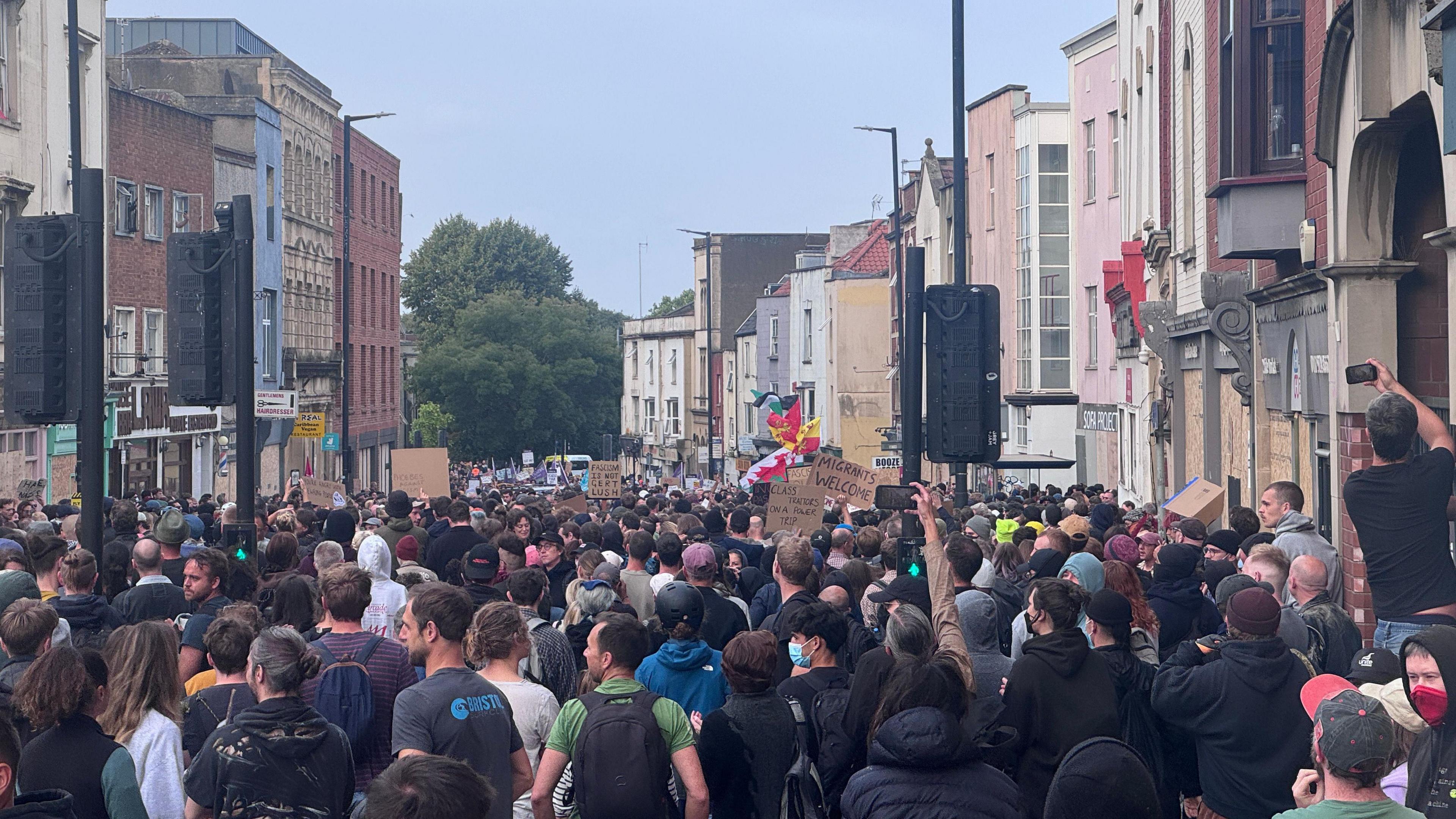 A huge crowd of people, some carrying cardboard signs or flag, walking down a city street. Most people are facing away from the camera and appear to be walking together. 