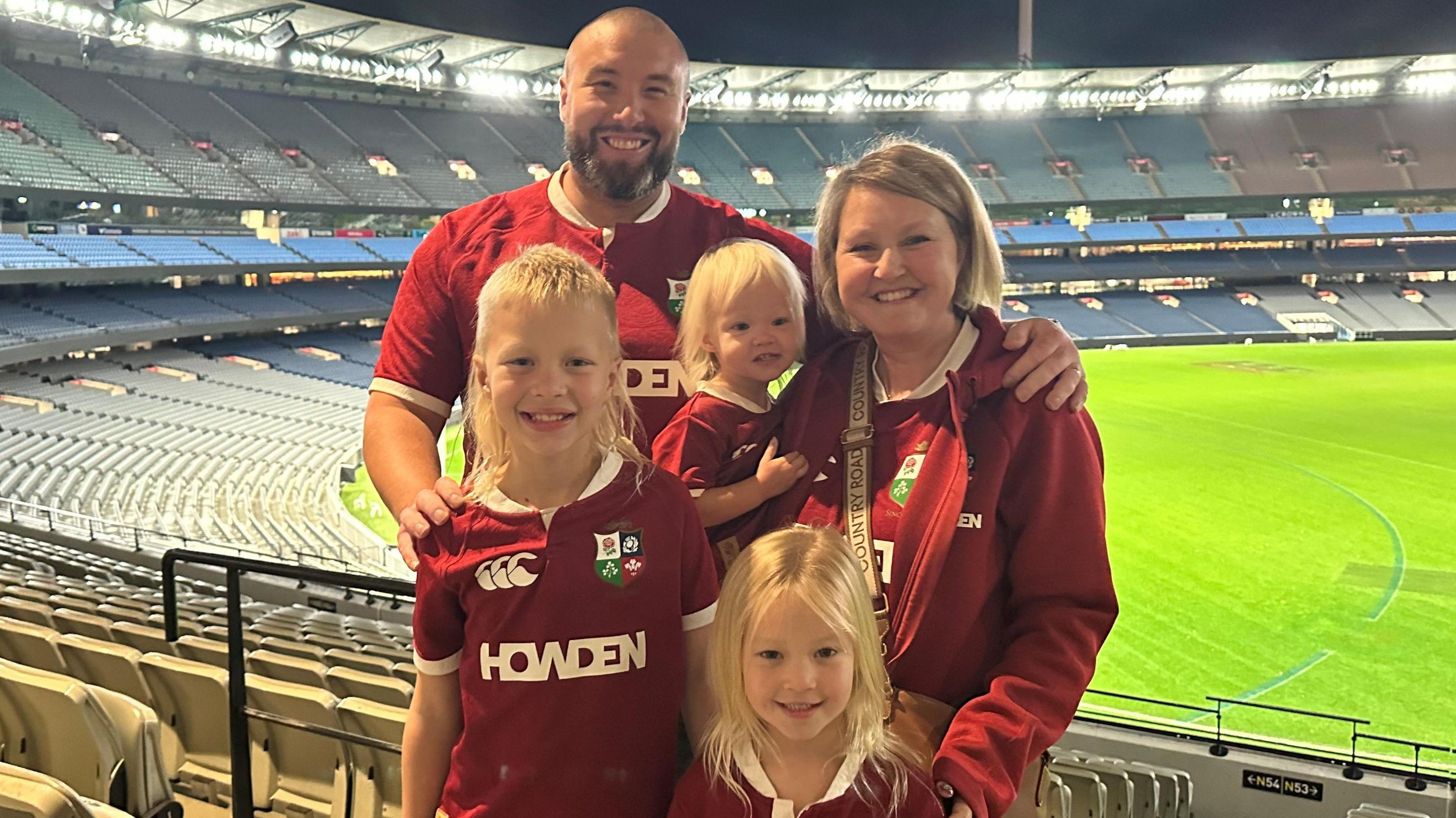 A family of a man and woman and three children in red British & Irish Lions shirts, standing in an empty stadium.
