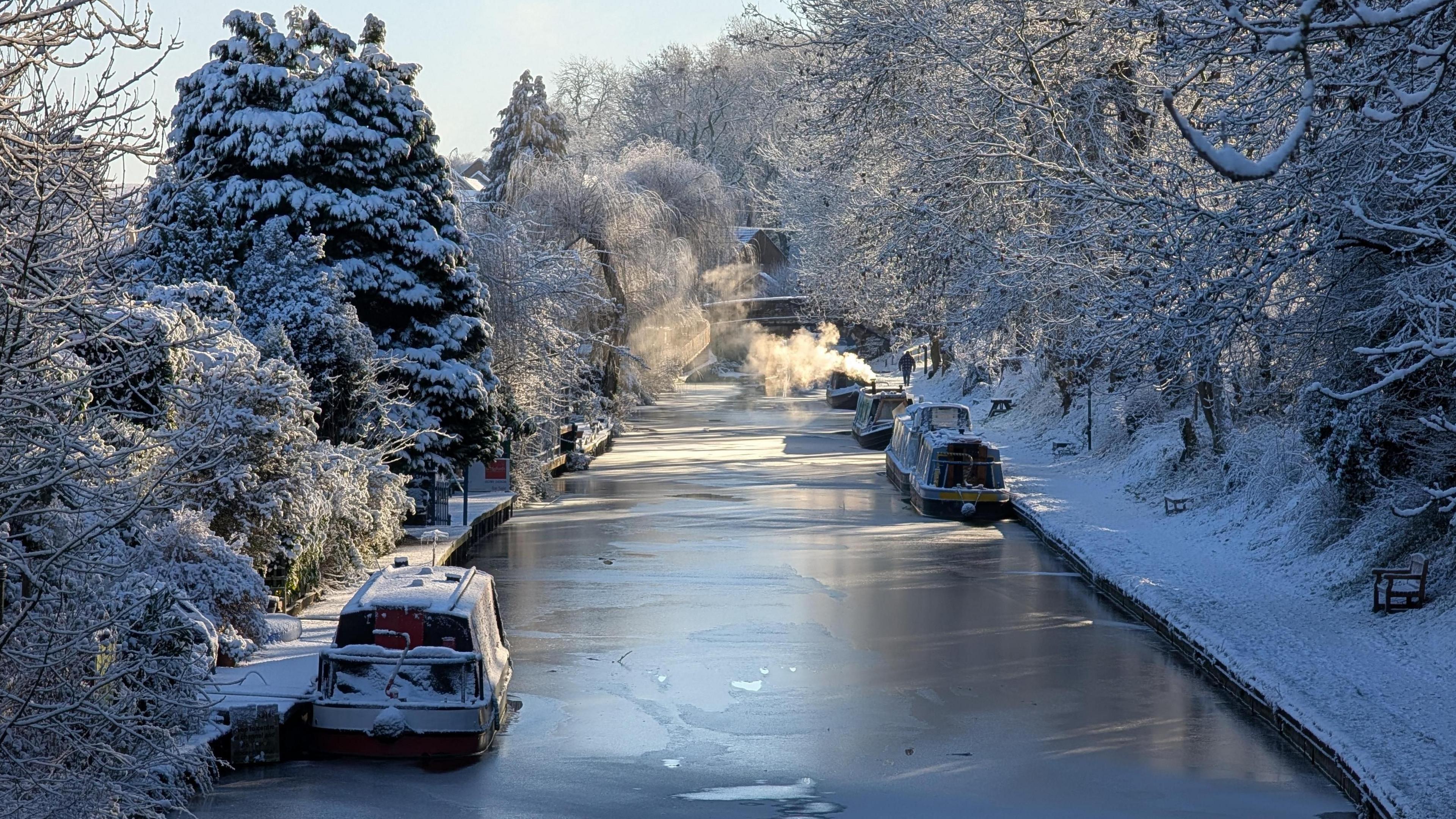 Canal flanked by snow-covered trees. Four barges float on the canal, which is covered in a layer of ice