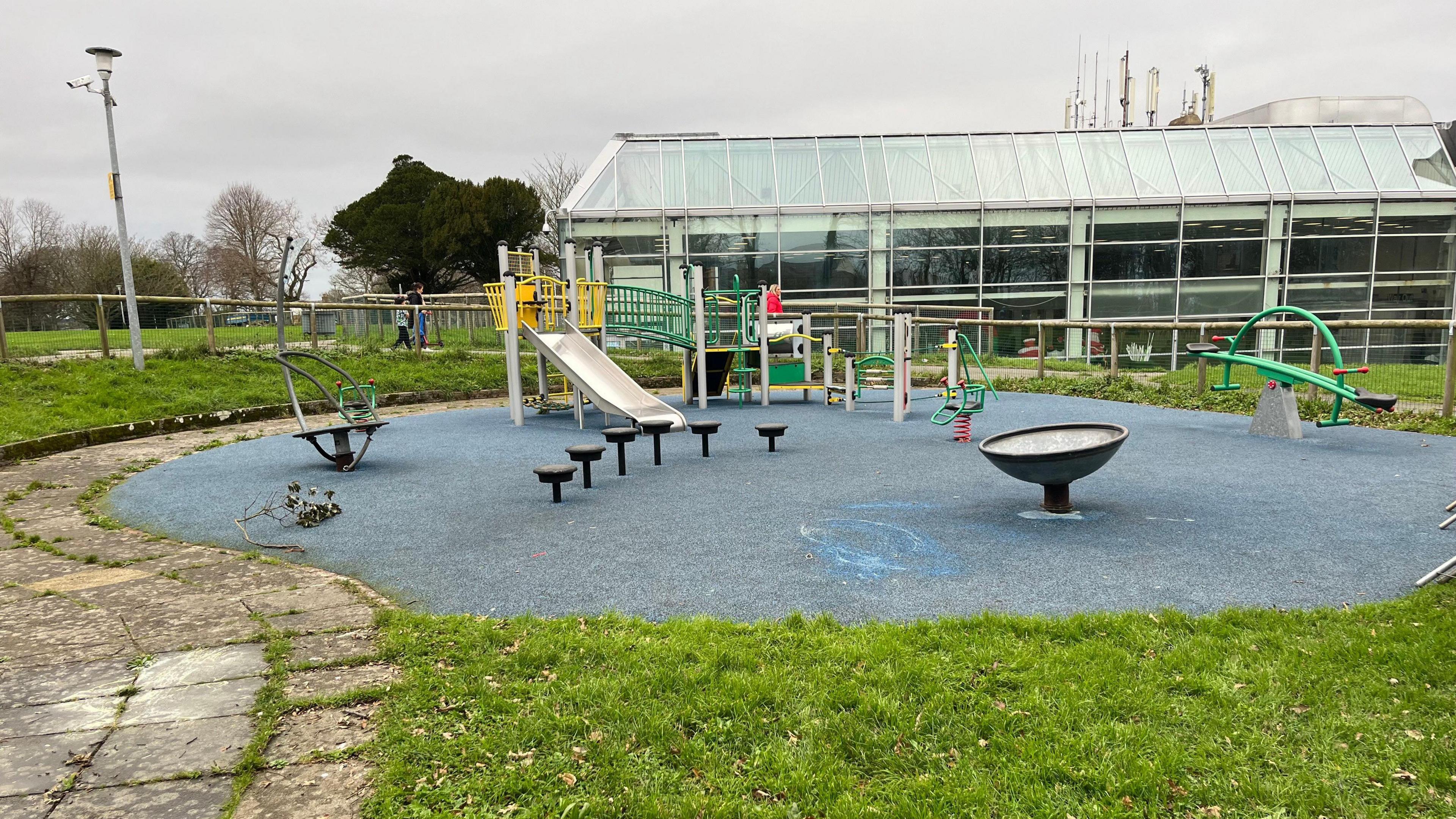 A view of Beau Sejour playground. There is blue matting on the floor which appears to have a swirled blue paint stain in one area. The play equipment includes a slide, a see saw and a climbing frame. The park is surrounded by grass and a stone path.