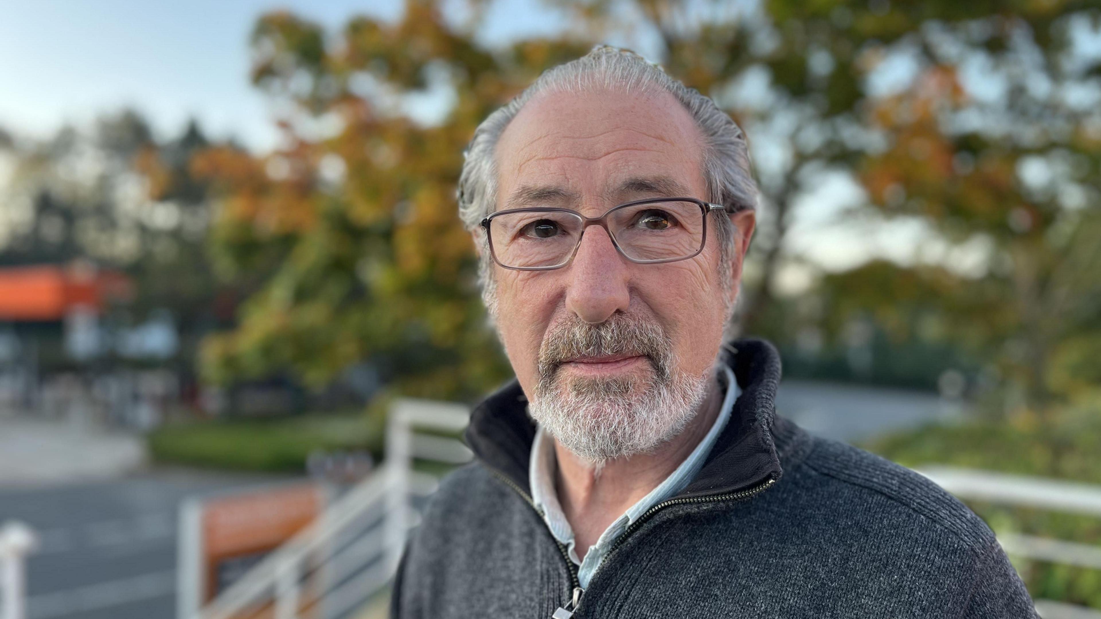 A man with grey hair and glasses wearing a grey jumper. There are some trees and a road out of focus in the background.