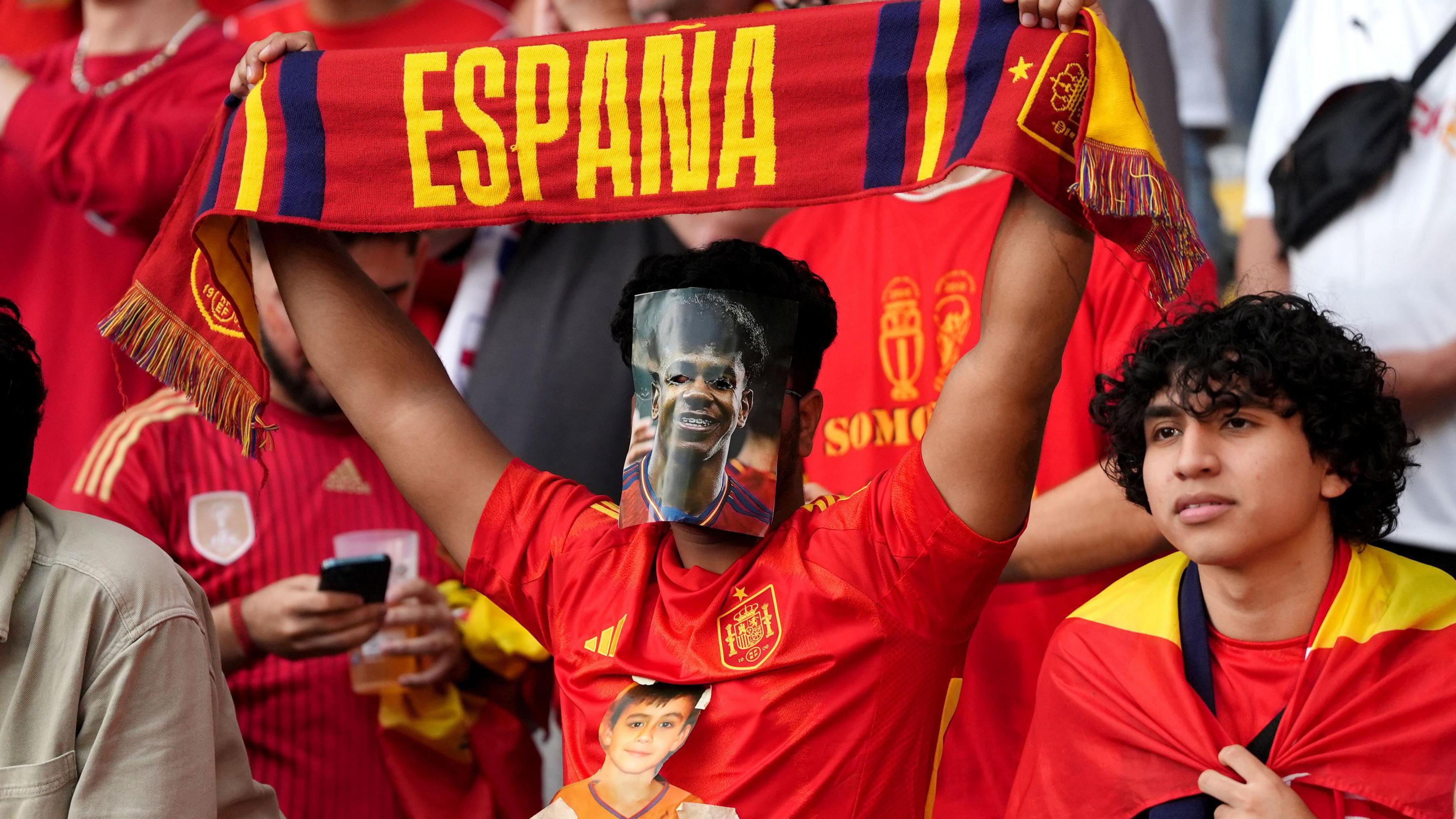 Spanish fan holds up a red and yellow scarf will wearing a mask of Lamine Yamal covering his face