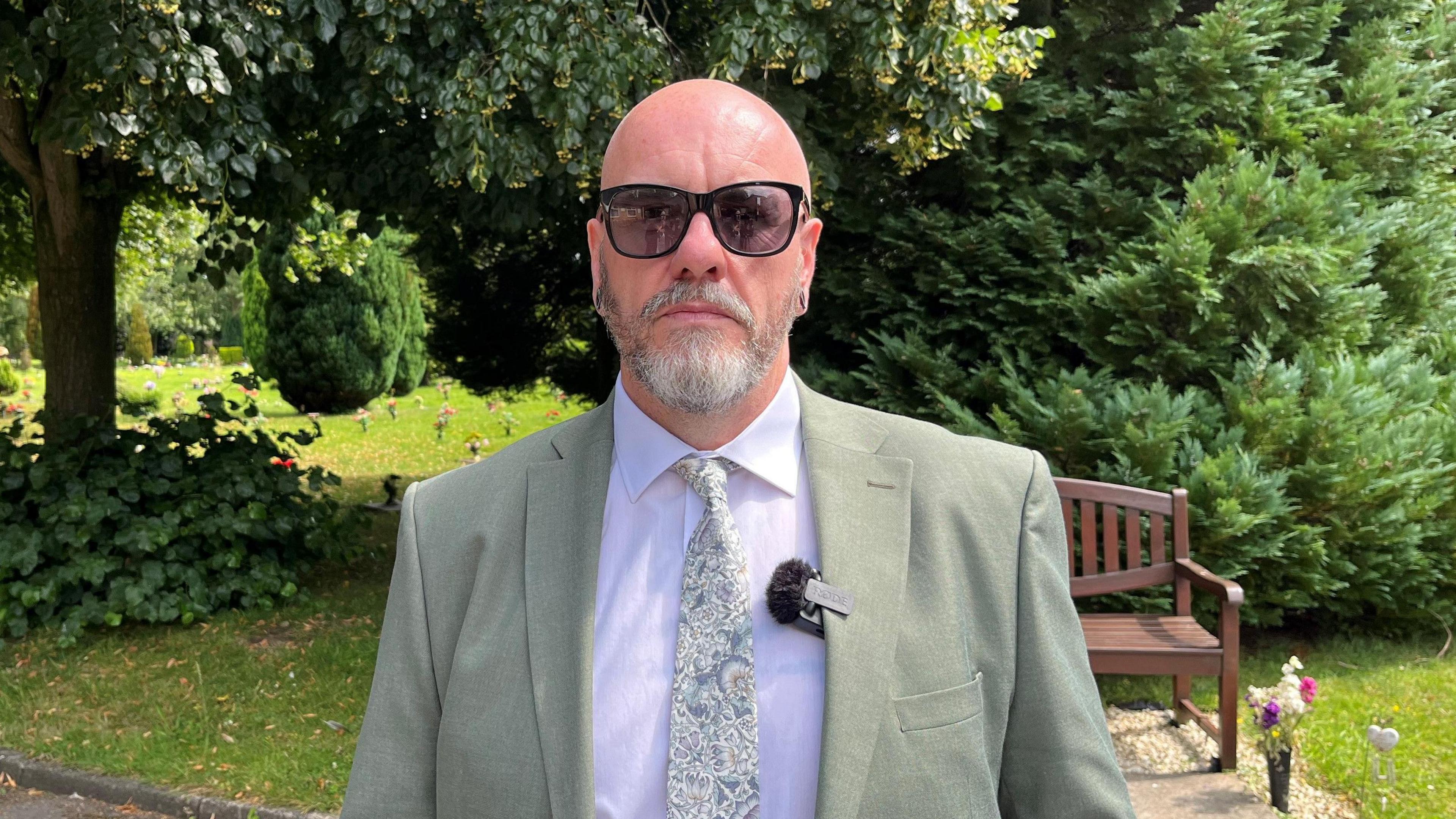 A man wearing sunglasses and a suit standing in front of a bench.