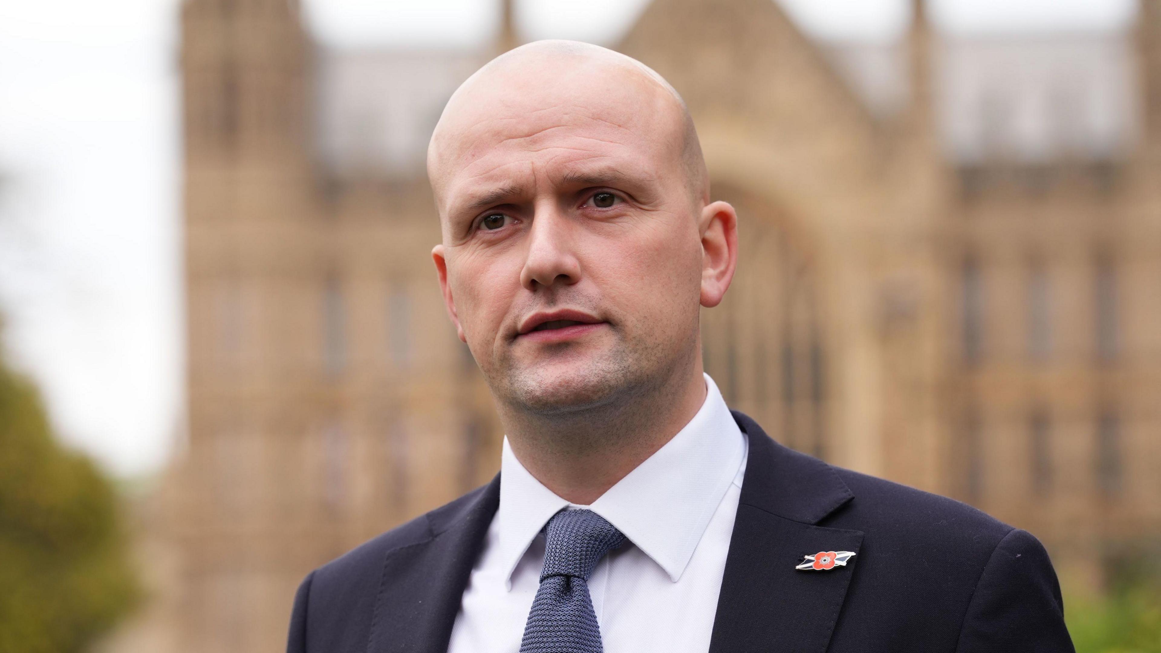 Stephen Flynn looking at the camera. The House of Commons is behind him. He is wearing a white shirt with a dark tie and a dark jacket with a poppy badge on the upper left lapel.