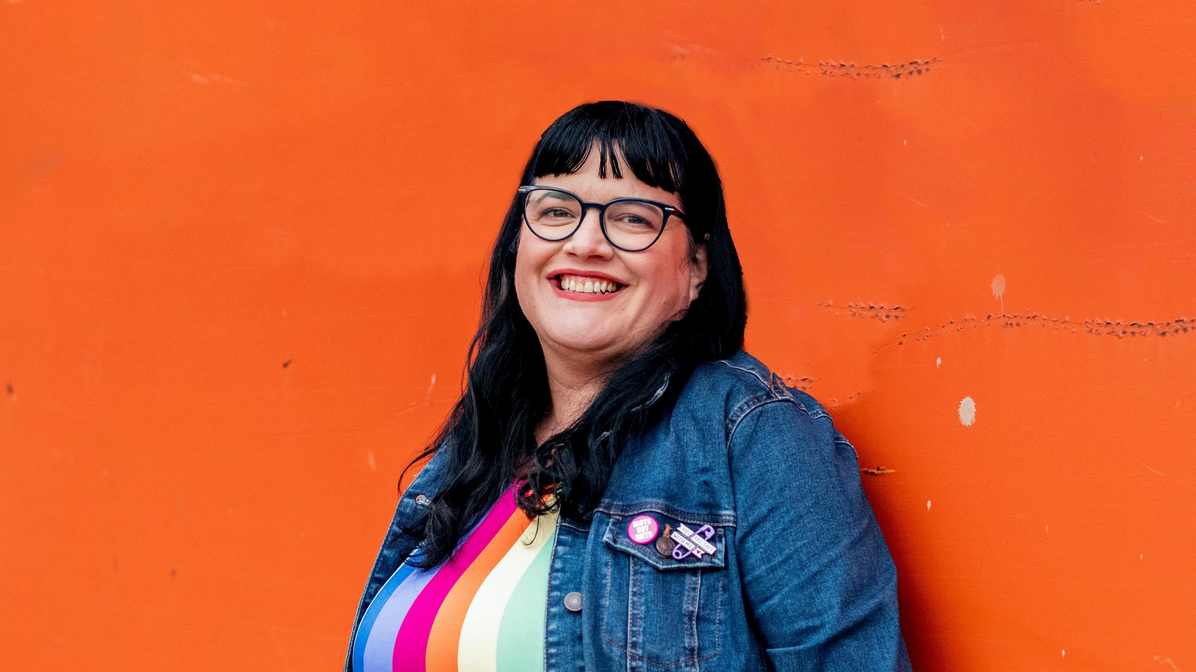 Heidi Mavir, wearing a stripy top and a jean jacket. She is standing in front of an orange wall smiling. She has long, black hair and is wearing thick-rimmed, black glasses.