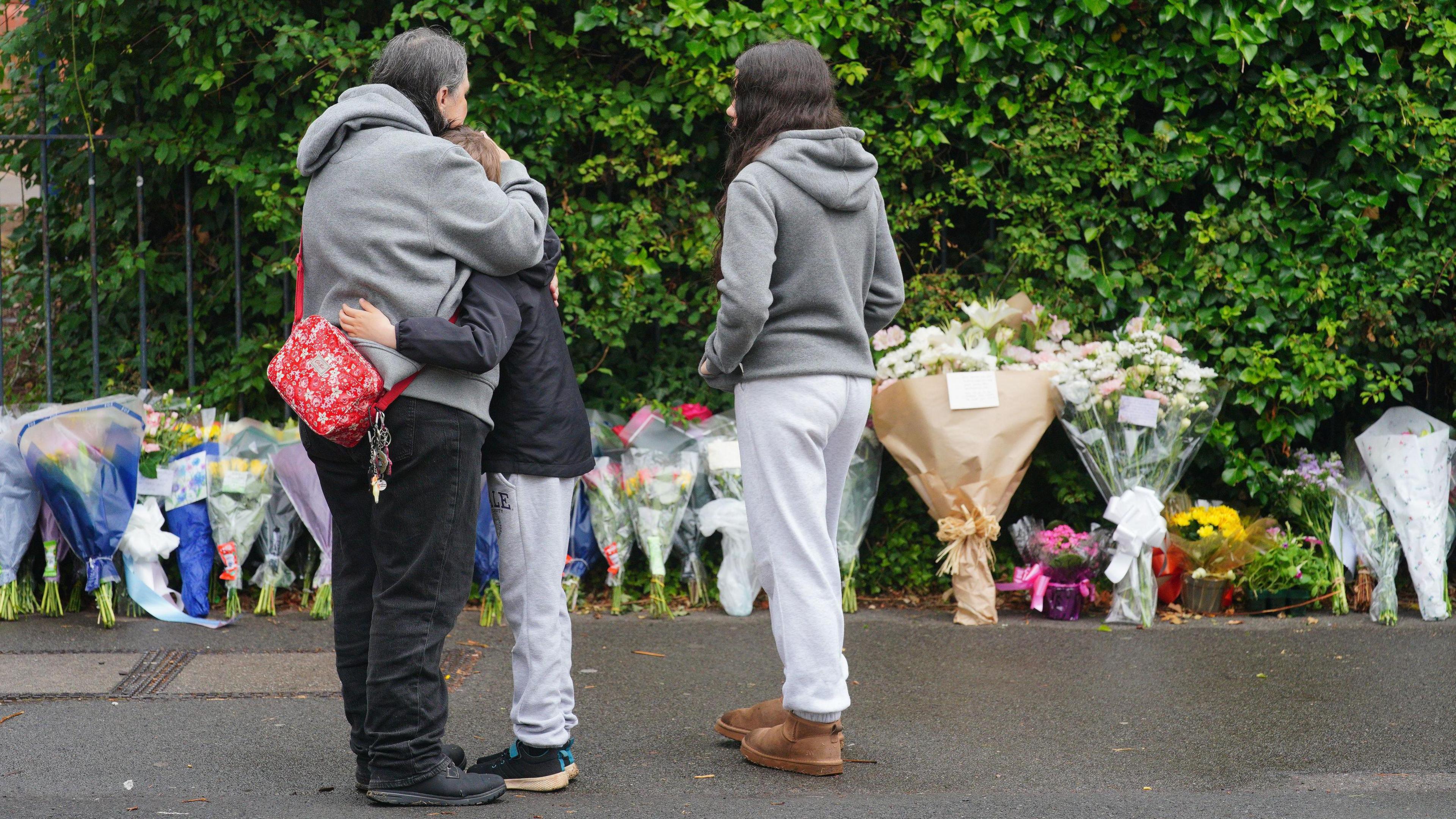 Three people standing outside Minehead Middle School. A woman is hugging a little boy and a young girl is stood next to them. There is a row of flowers left in tribute to a pupil who died in a coach crash. 