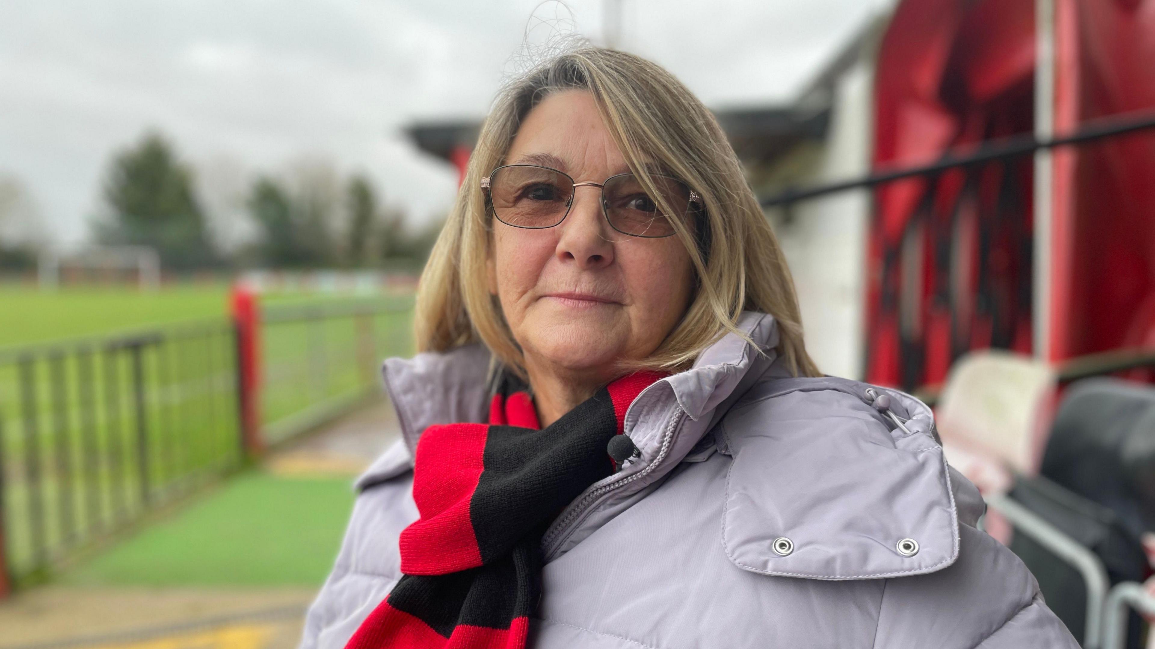 Pat Boyd, wearing glasses, a light lilac coat and a red and black scarf. She is sitting in a football grandstand.