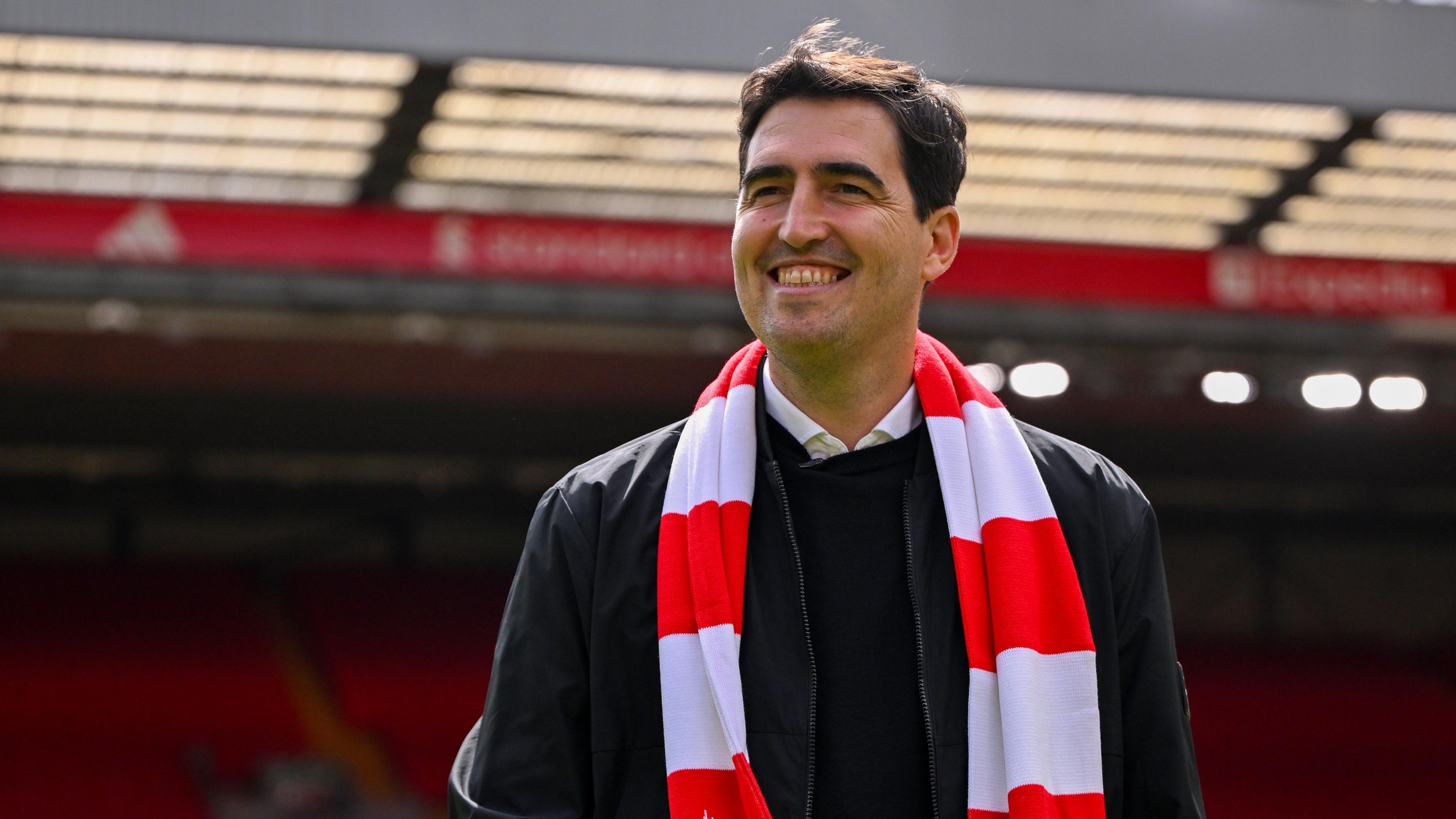 Andoni Iraola smiles while standing on Anfield pitch wearing a Liverpool red and white scarf
