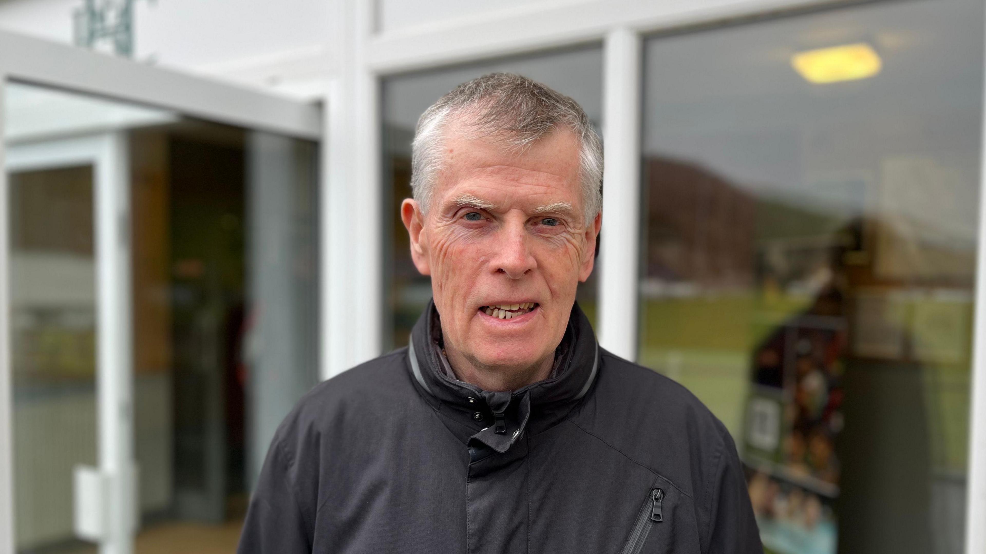 A grey-haired man with a charcoal zipped up jacked look at the camera