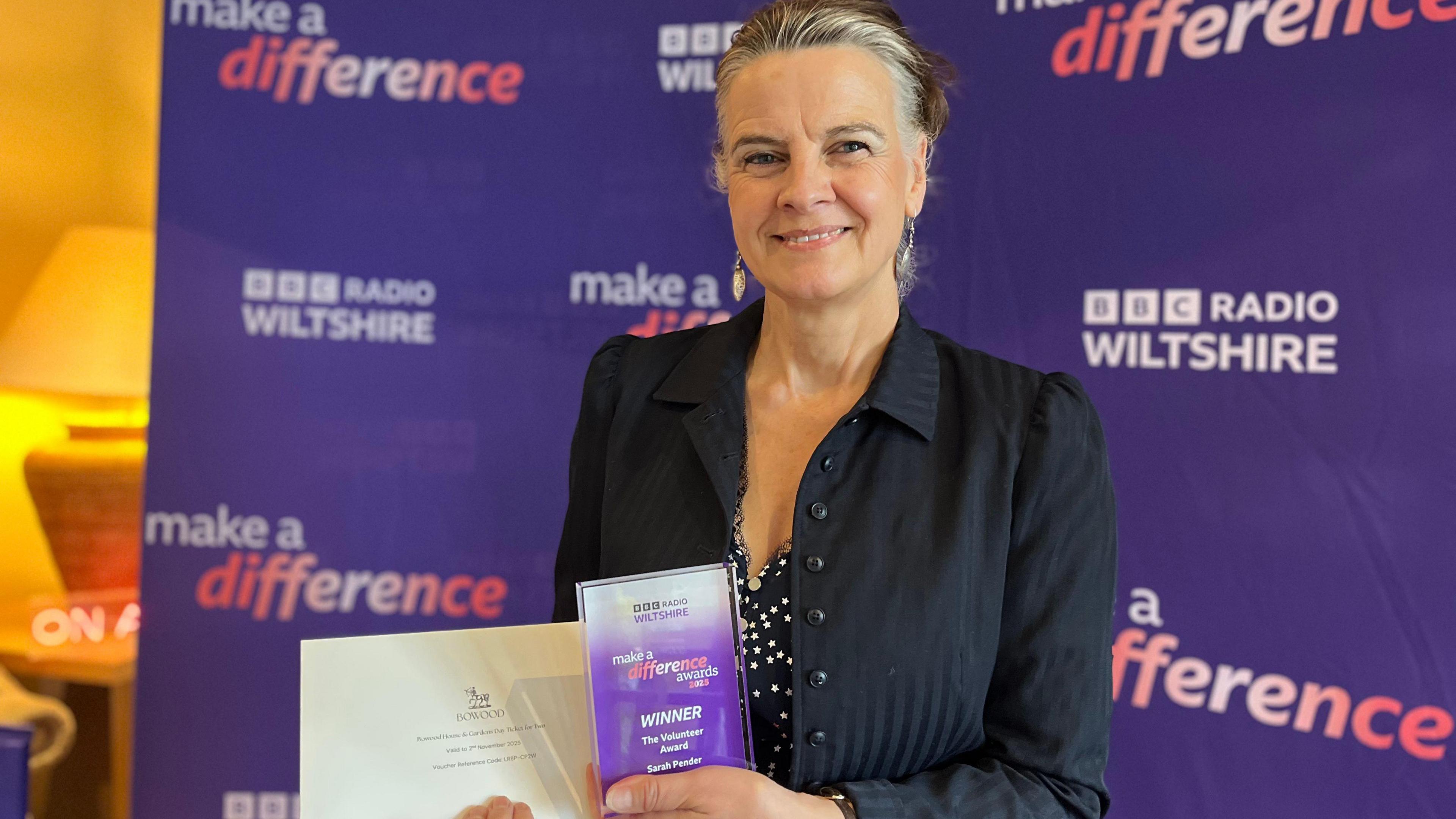 Sarah Pender smiles at the camera holding her award. She is wearing a navy blue jacket and polka dot dress. Behind them is a purple banner saying BBC Radio Wiltshire and Make a Difference.