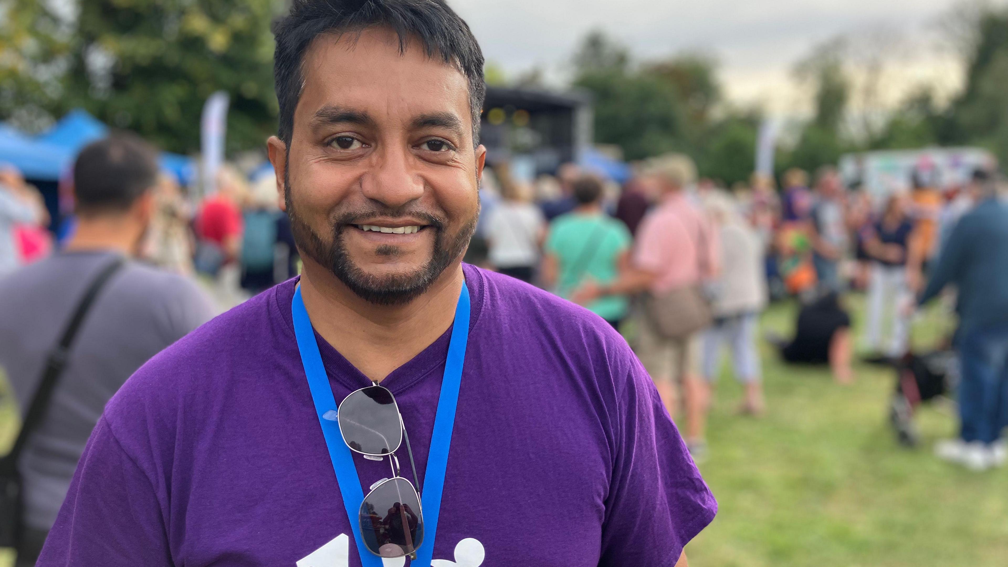 Boshor Ali stands in a park in a purple t-shirt with crowds of people behind him.