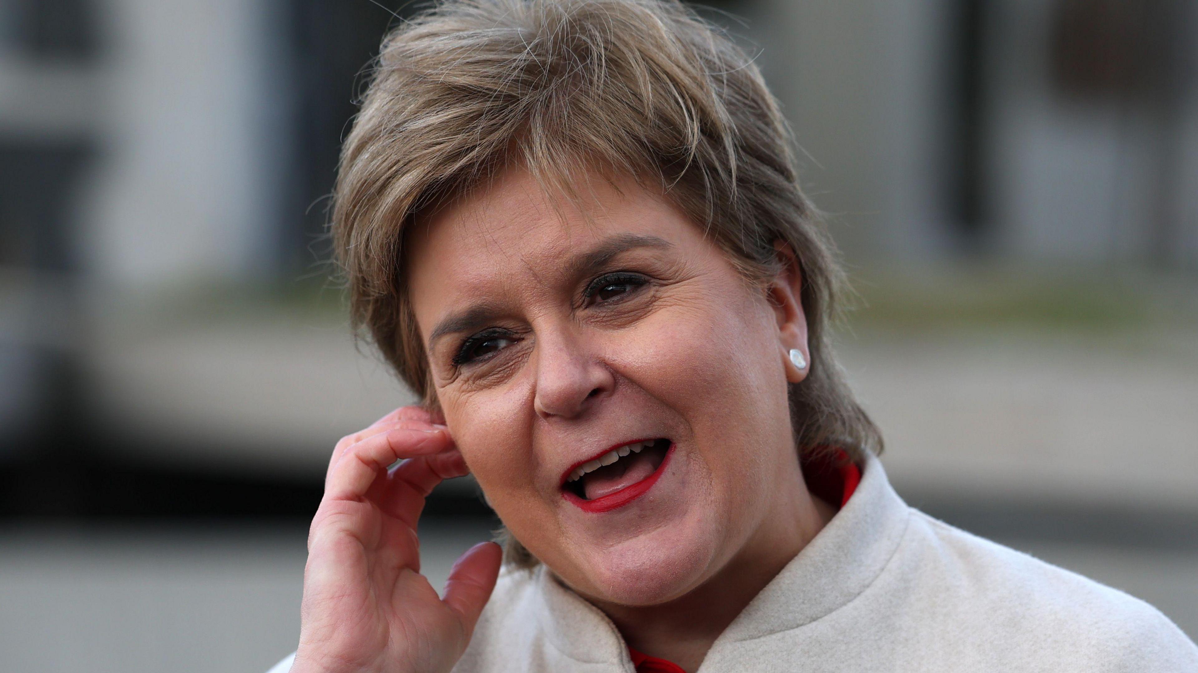 Headshot of Nicola Sturgeon touching an TV earpiece in her right ear. She has short brown/ blonde hair and is laughing. She is wearing a beige jacket.