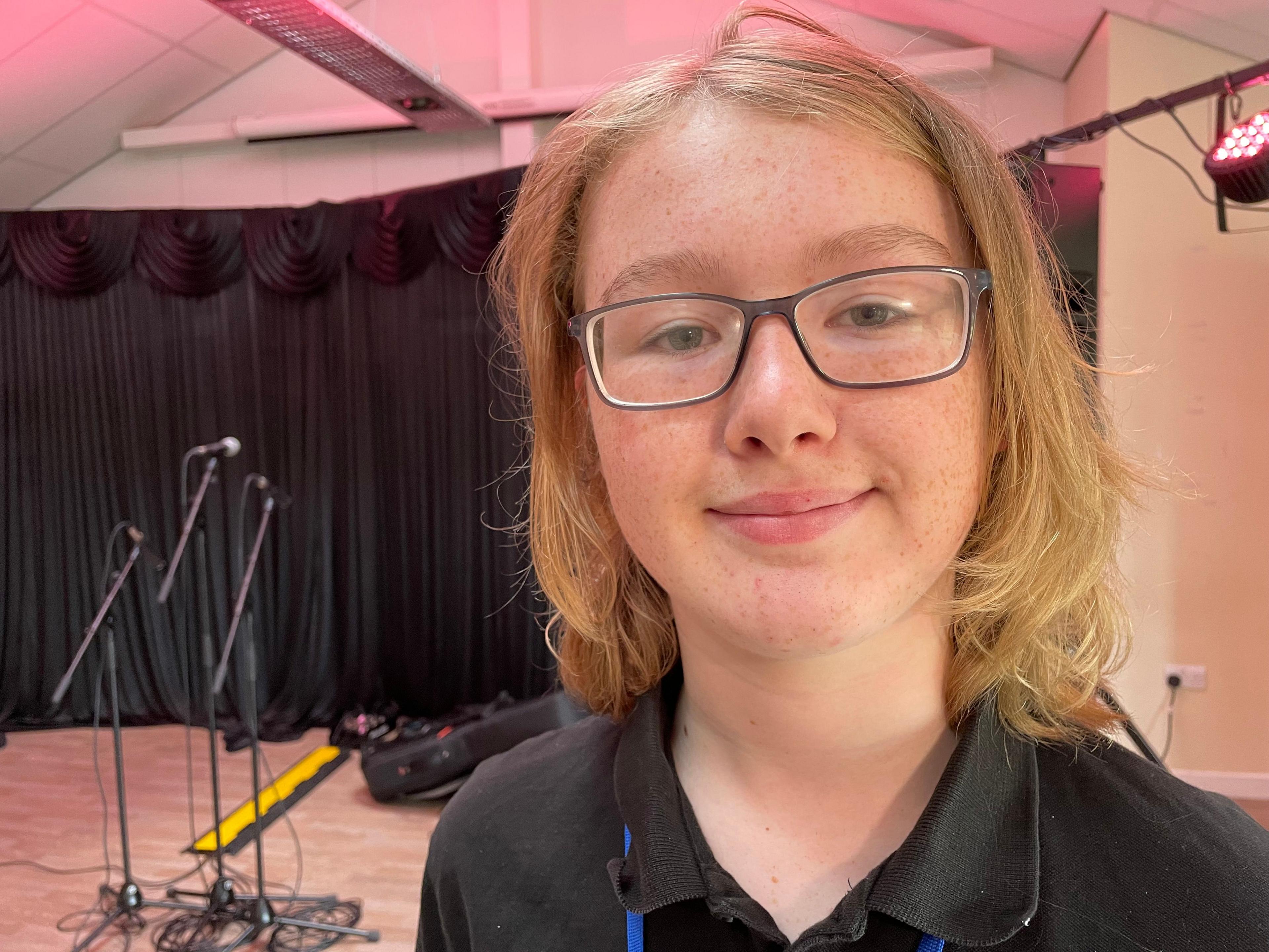 A teenage boy with shoulder-length blonde hair and glasses.