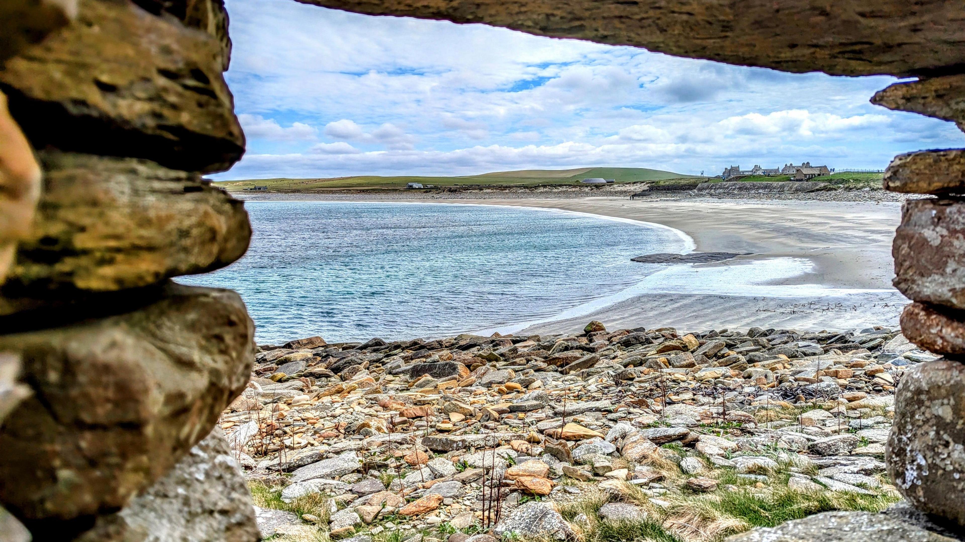 Skaill Bay on Orkney. There is a beach on the right side of the image. The water on the left side of the image is blue. The sky is a lighter blue and is streaked with clouds. In the foreground, there is multi-coloured shale in orange, brown and light colours. The entire image is framed by a stone window.