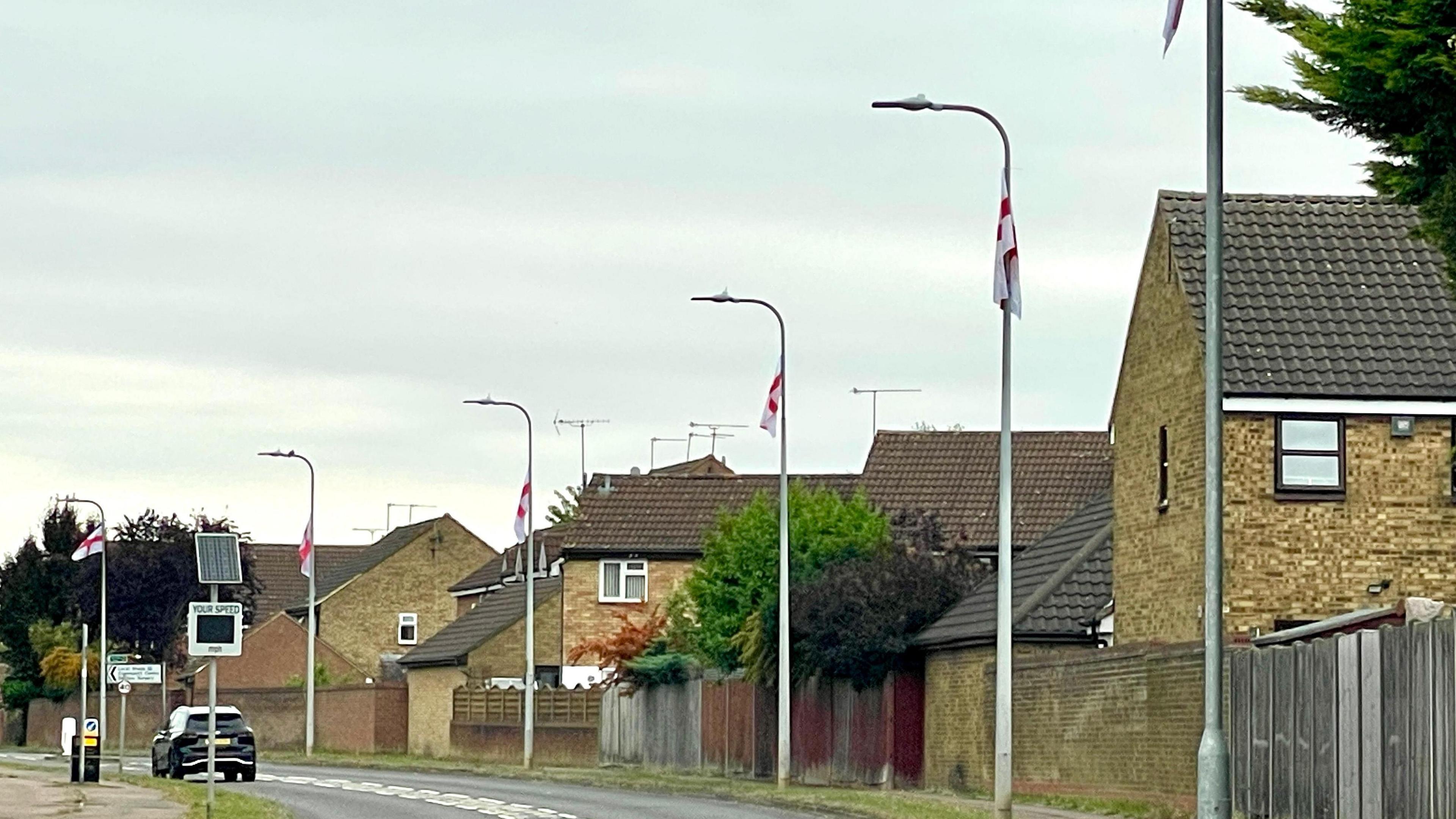 A row of six lamp-posts evenly spaced out next to housing. England flags have been attached to the tops of the straight parts of the posts, just under where they curve out over the road.