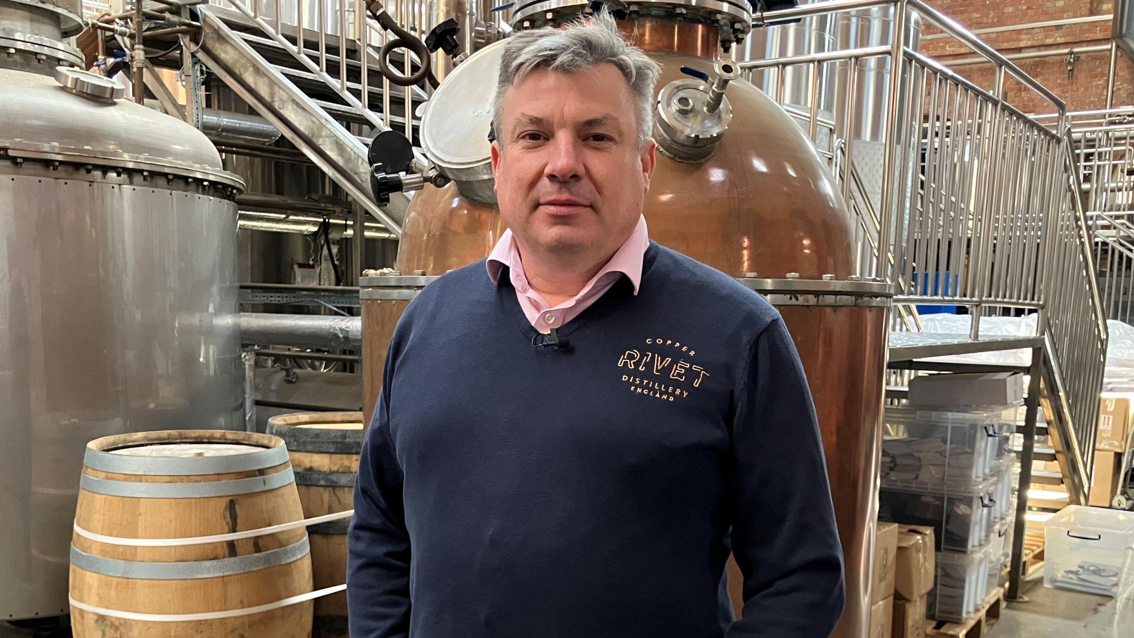 The co-founder of the Copper Rivet Distillery in Chatham, Matthew Russell, pictured in the distillery. In the background there are several copper pots for making the gin and several barrels for storing it. Matthew has greying hair and is wearing a navy branded jumper and pink undershirt.